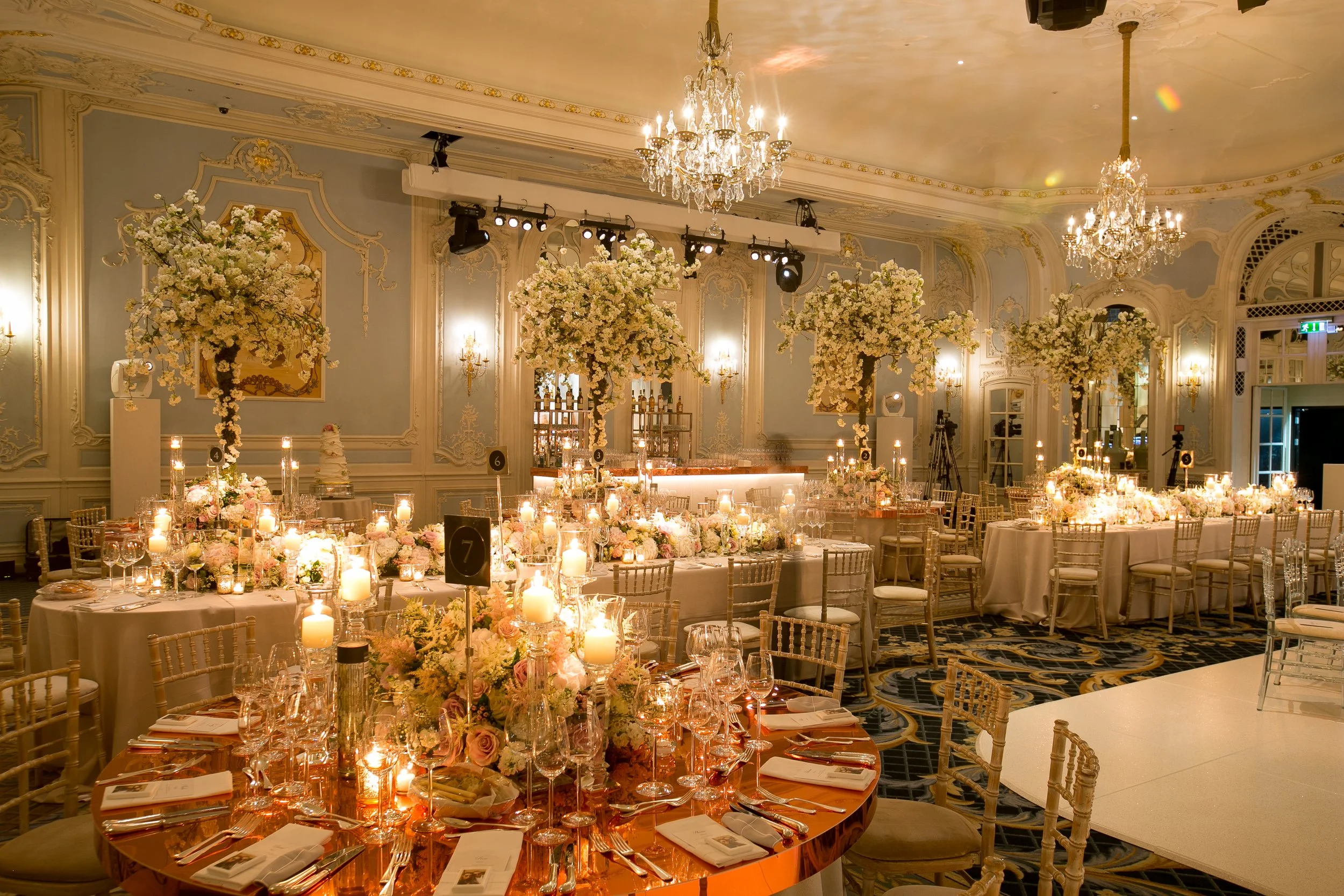 Elegant banquet hall decorated with large floral centerpieces, numerous lit candles, and chandeliers, set for a formal event.