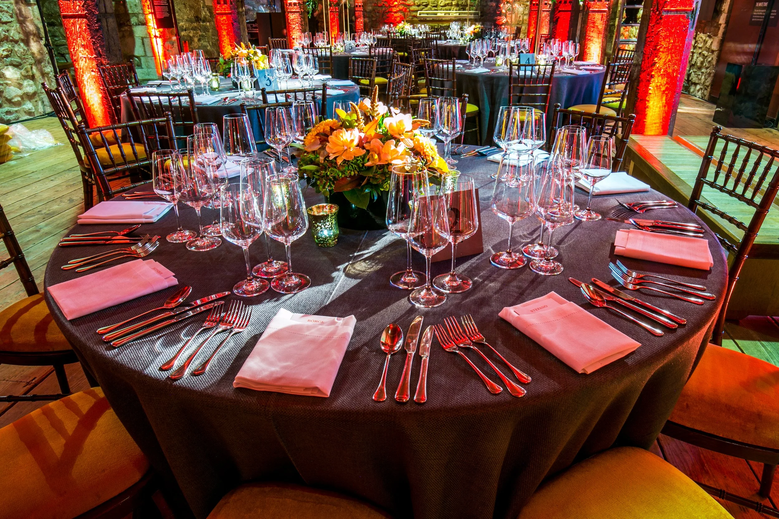 Elegant round dining table set for a formal dinner with multiple wine glasses, silverware, white napkins, and floral centerpiece in a warmly lit venue with exposed stone walls and wooden flooring at the Tower of London