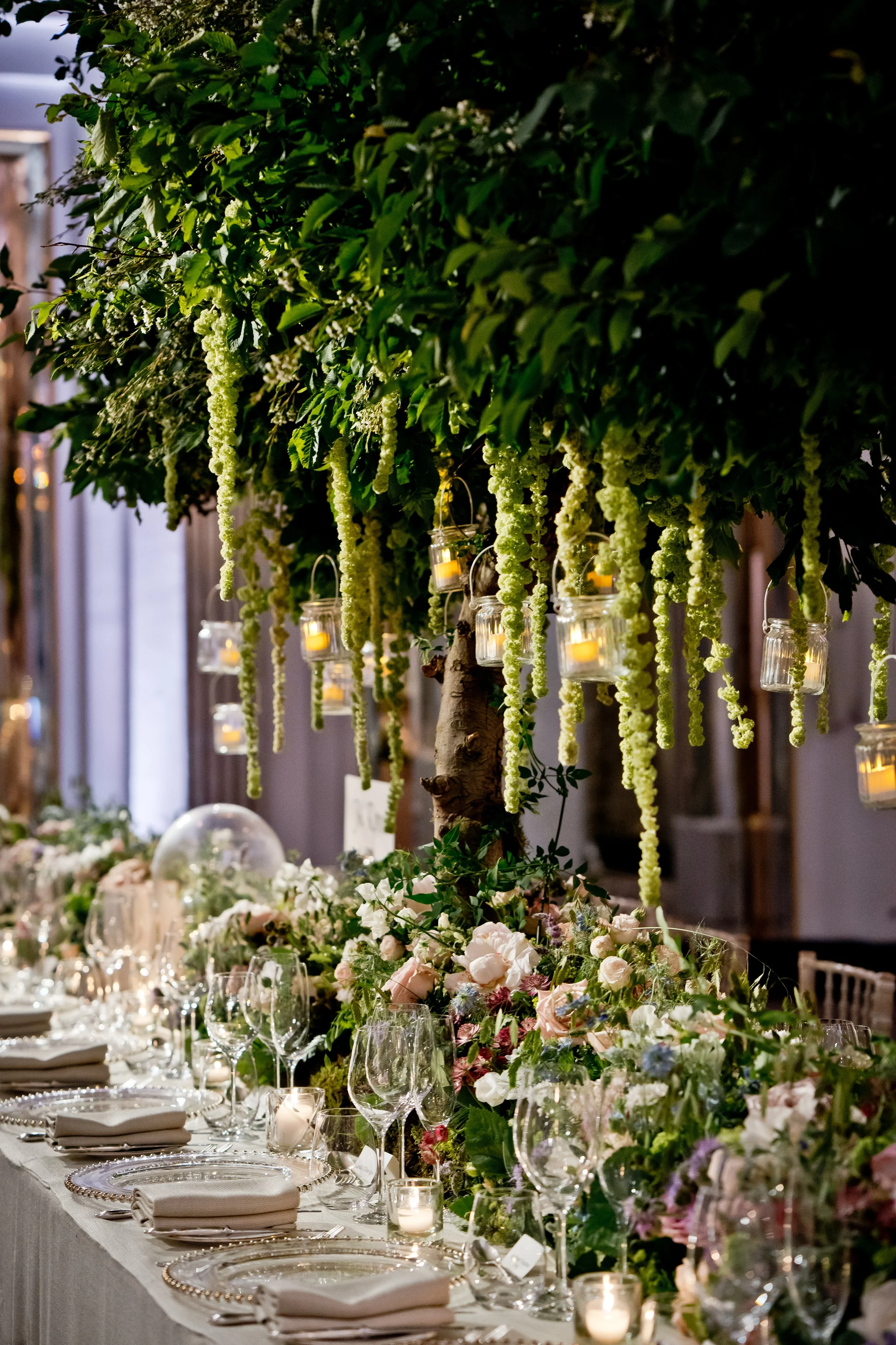 Elegant banquet table decorated with a lush floral arrangement of white and pastel flowers and greenery, with hanging glass lanterns containing candles, set for a formal event.