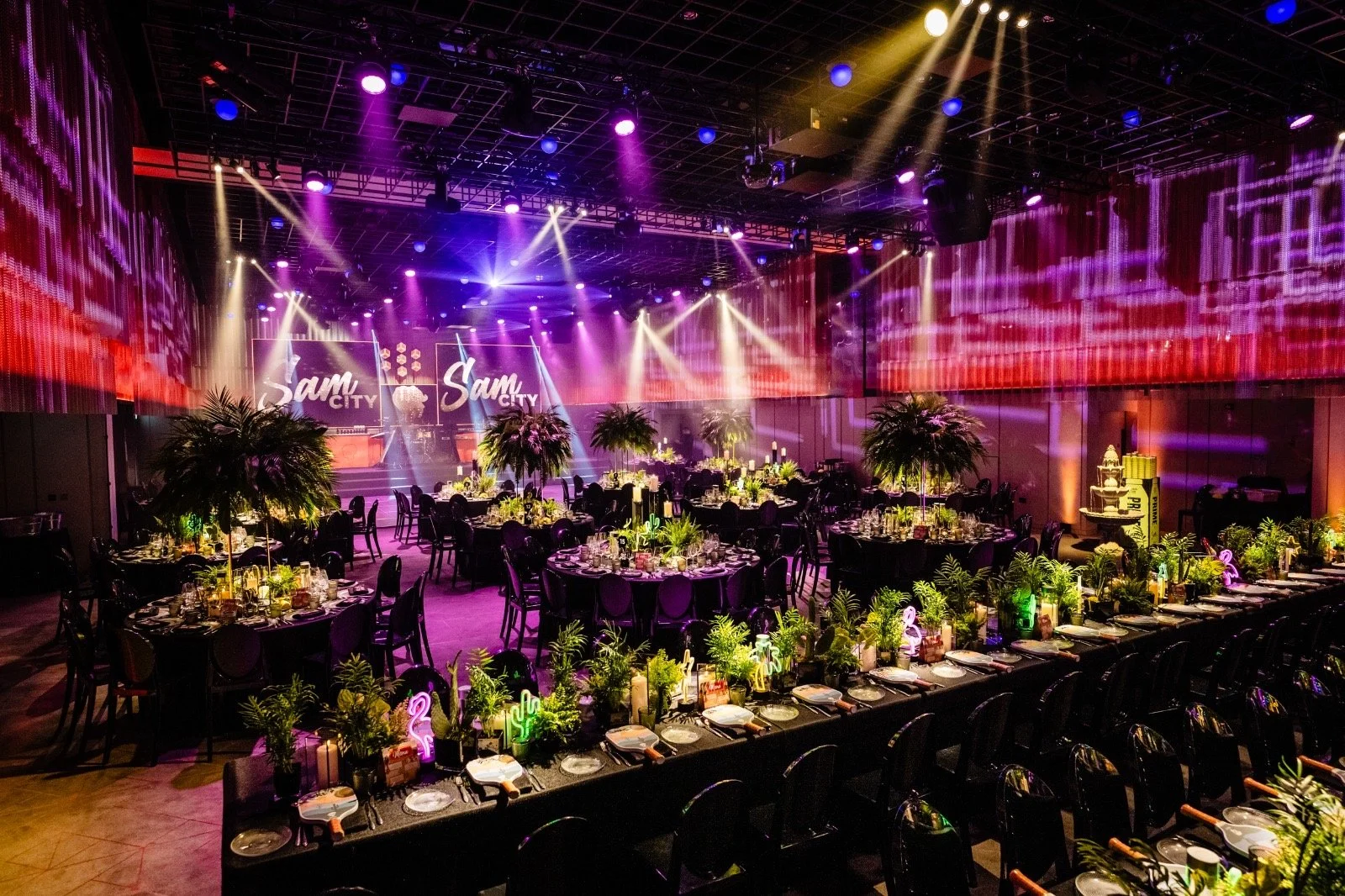 A decorated event space with round tables set with plates, glasses, and silverware, surrounded by black chairs. The tables have centerpieces with green plants and candles. Large leafy plants are also on the floor. The background features a stage with two large screens displaying "Sam City," and colorful lighting with purple, pink, and yellow beams illuminate the room, creating a lively atmosphere.