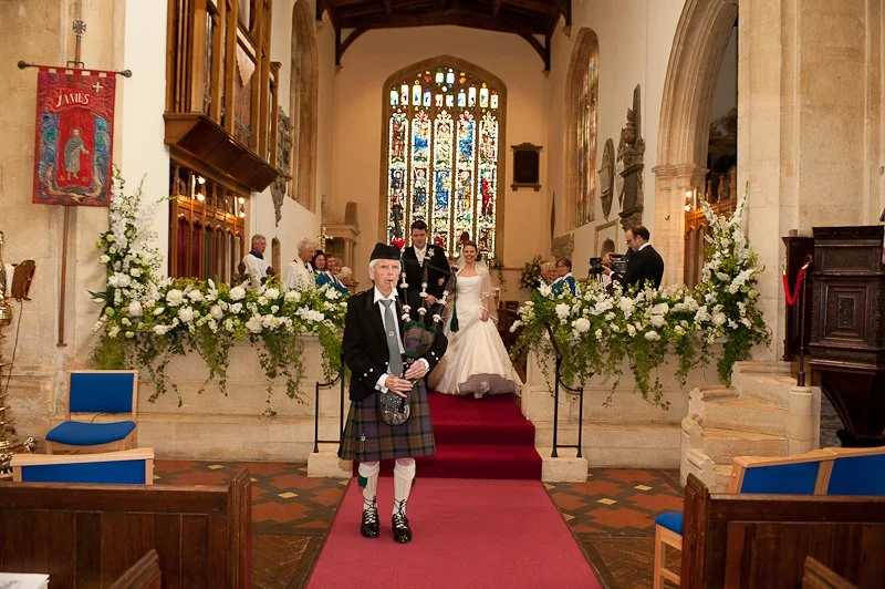A wedding ceremony inside a church with a bride in a white wedding dress, a groom, and guests. A bagpiper in traditional Scottish attire is playing in the foreground. The church has stained glass windows and floral decorations.