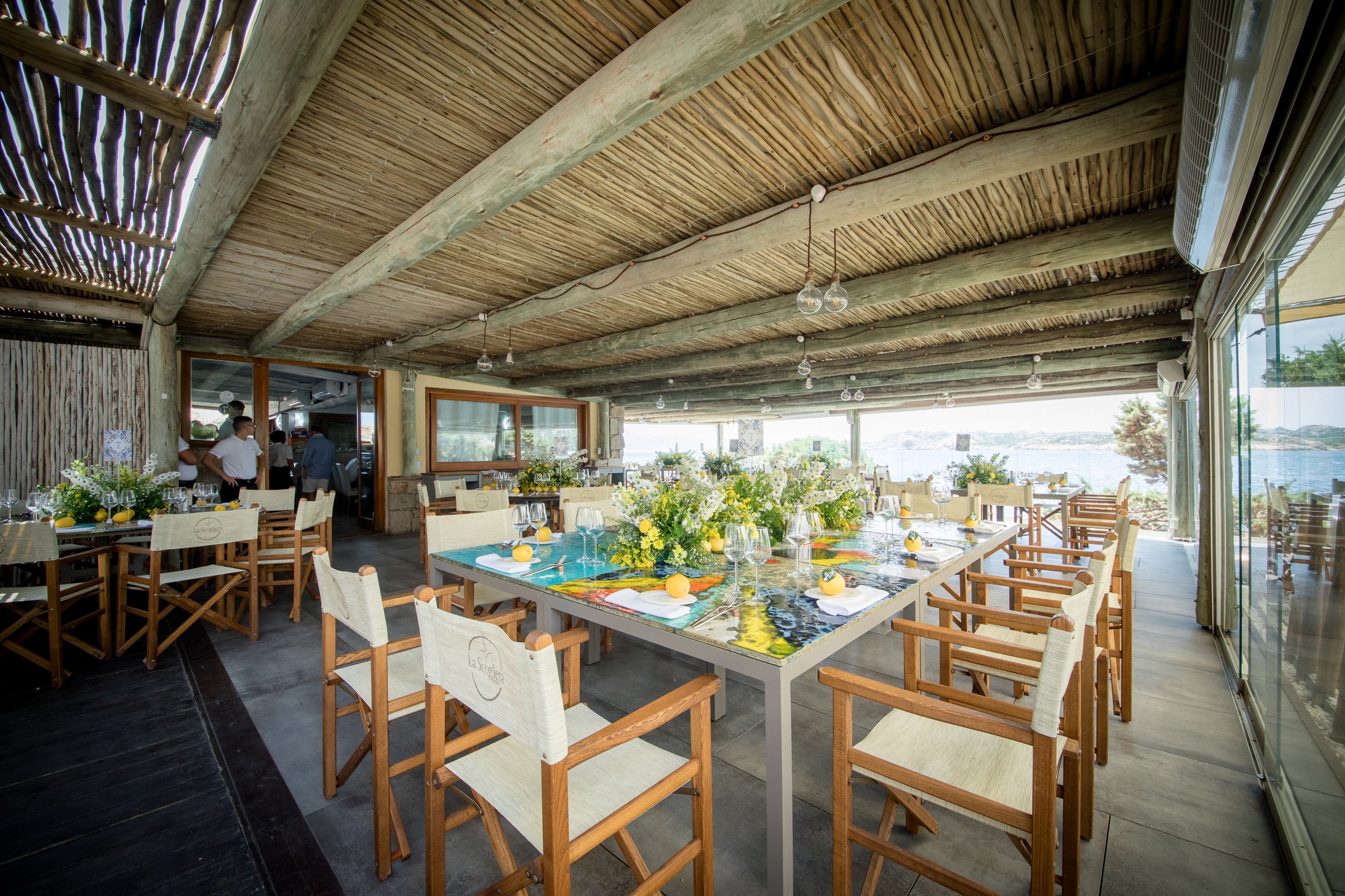 A dining area with long tables decorated with yellow and white flower arrangements and lemons, set with wine glasses and white napkins. The space features a wooden ceiling with exposed beams and large windows providing a view of water and greenery ou