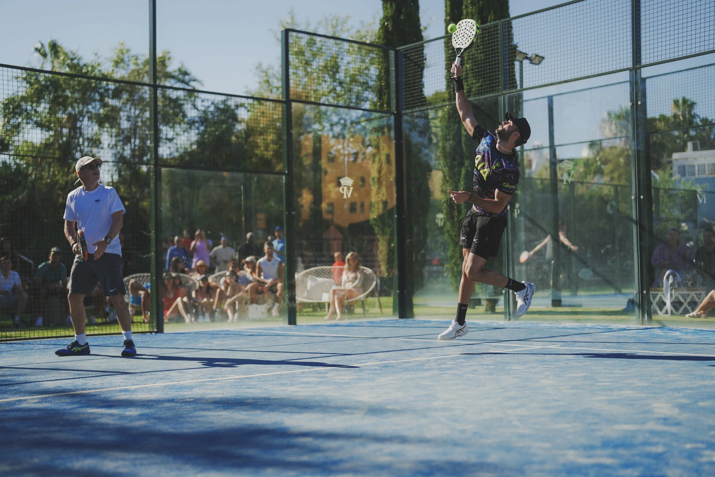 A young man plays a game of pickleball on an outdoor court, hitting the ball with a paddle while an older man watches, surrounded by spectators.