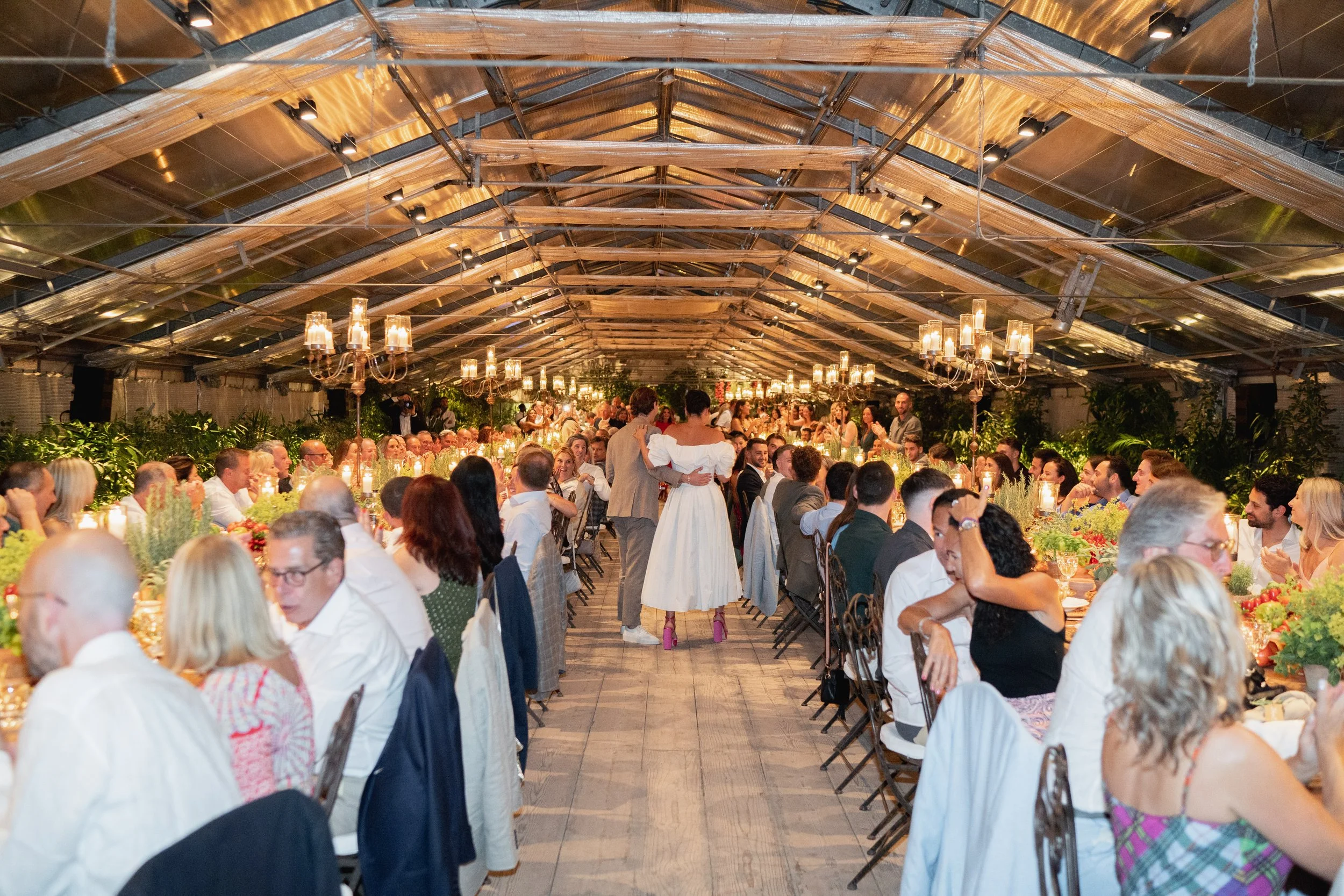 Crowd of people at a wedding reception or banquet, with a couple dancing in the center, under a wooden and glass ceiling with chandeliers and greenery decor. Venue is in Tuscany at Serre Torrigiani Bistrot