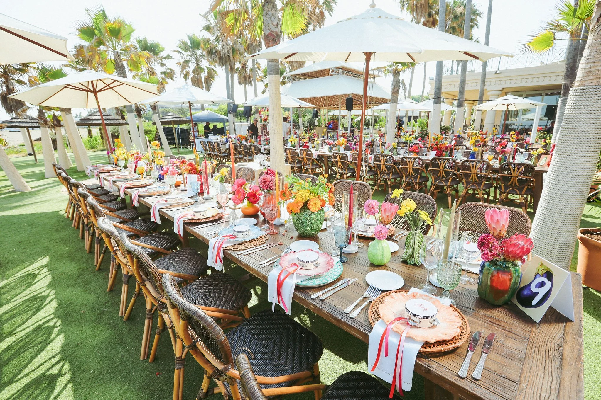 An outdoor dining setup with a long wooden table decorated with colorful flowers, glassware, and plates, surrounded by wicker chairs. Large white umbrellas, palm trees in the background in Marbella at Anantara Villa Padierna Resort 