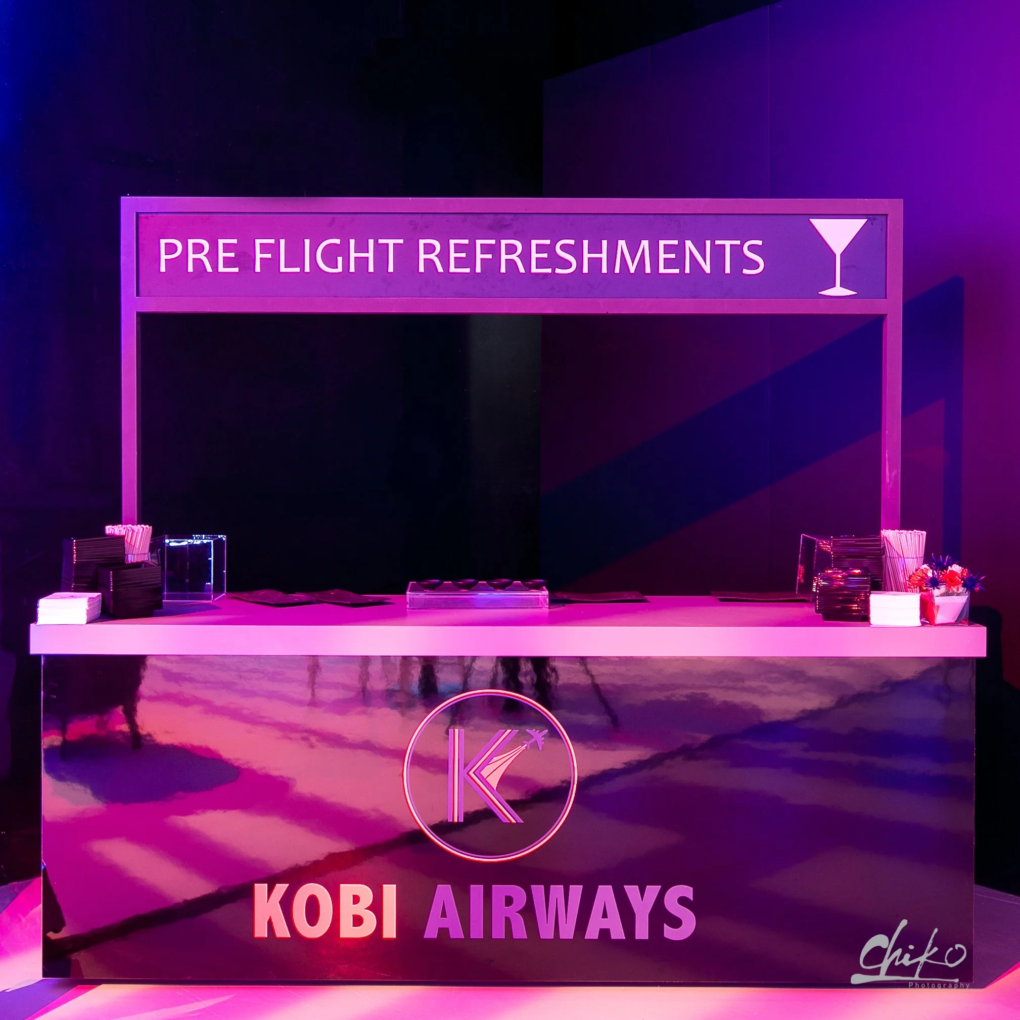 Neon sign reads 'Pre Flight Refreshments' with a martini glass icon, set against a dark background with purple and pink lighting at Kobi Airways