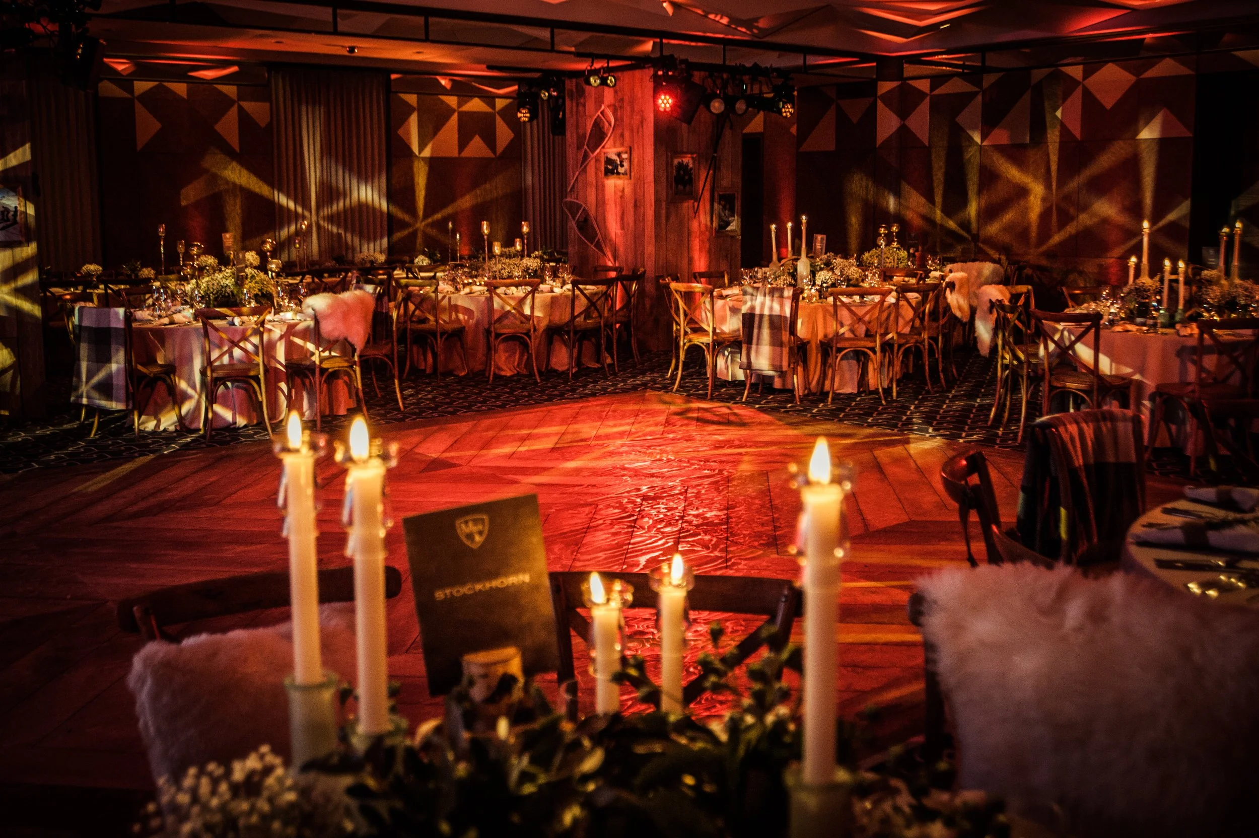 Elegant banquet hall decorated for an event with warm lighting, round tables with floral centerpieces, candles, and chairs with plaid cushions. Soft lighting creates a cozy atmosphere.