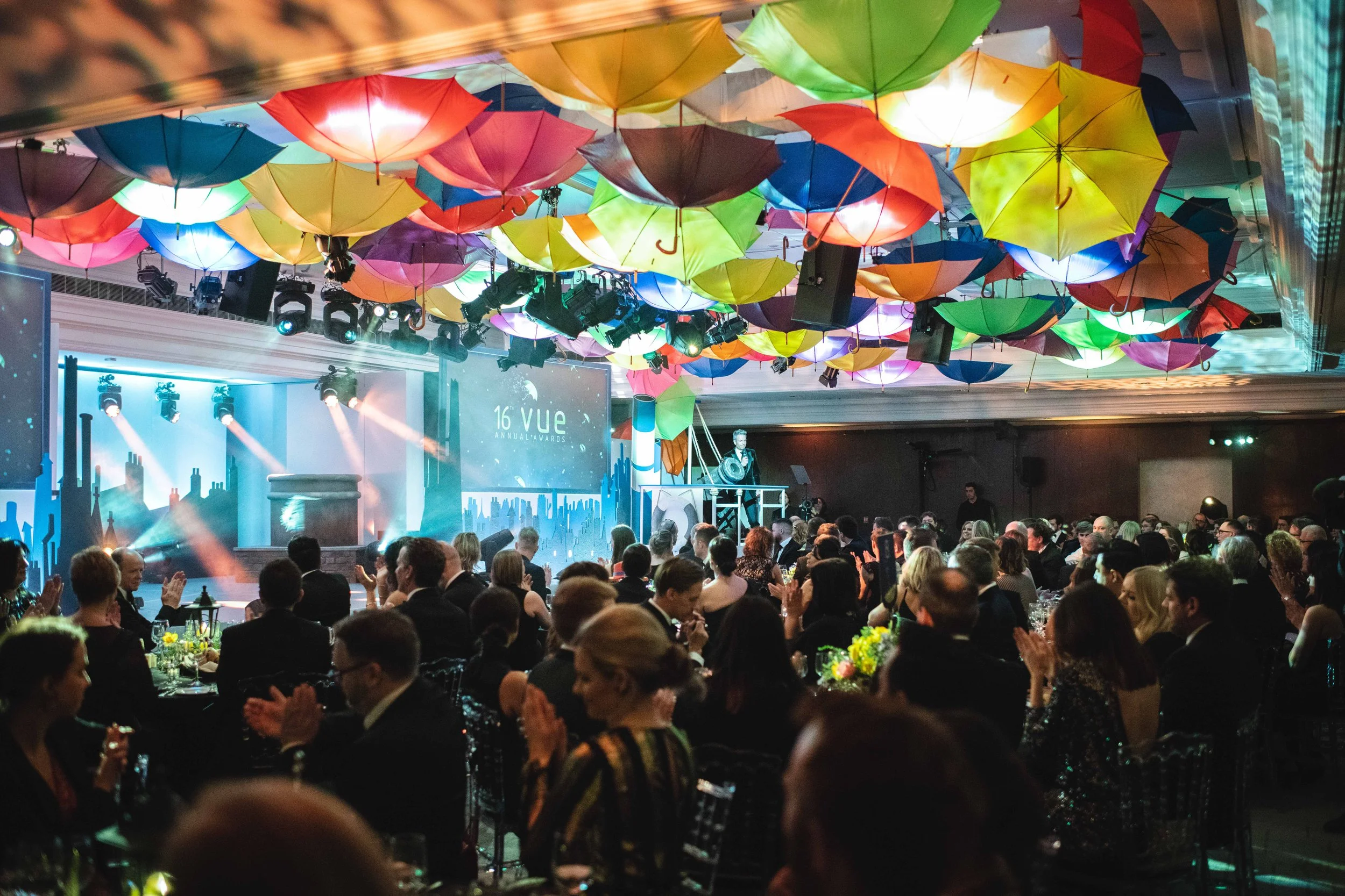 Large gathering of people at an indoor event with colorful umbrellas hanging from the ceiling. There is stage lighting, a speaker on stage, and an audience clapping and seated at tables with floral centerpieces.