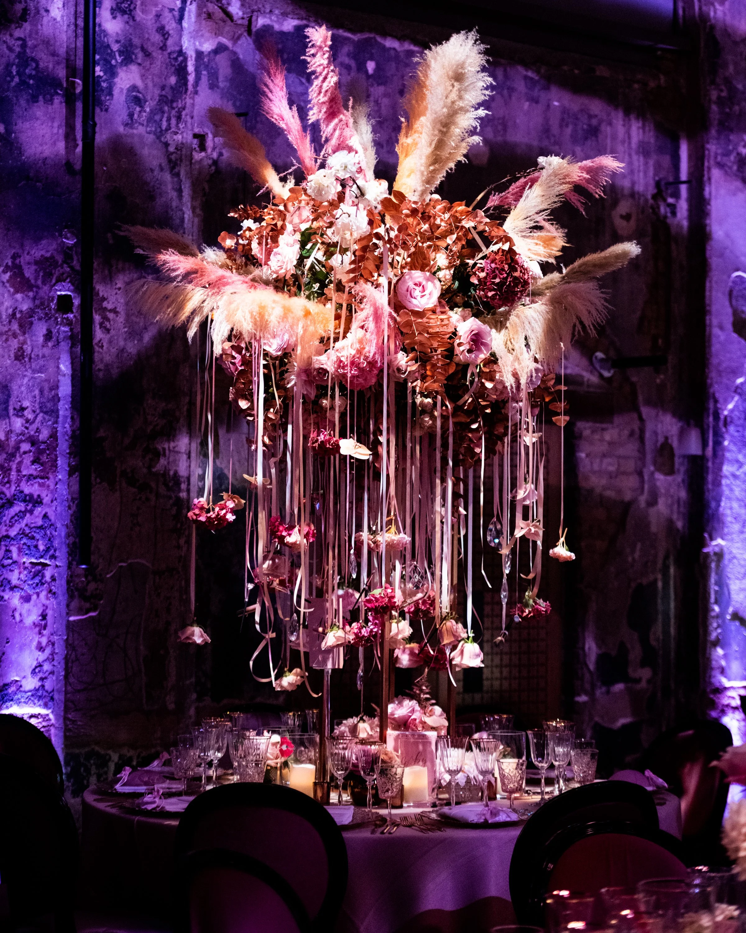 A large floral centerpiece with pink and white flowers, pampas grass, and hanging ribbons on a decorated banquet table, illuminated by pink and purple lighting against a stone wall. BAT MITZVAH AT BATTERSEA ARTS CENTRE