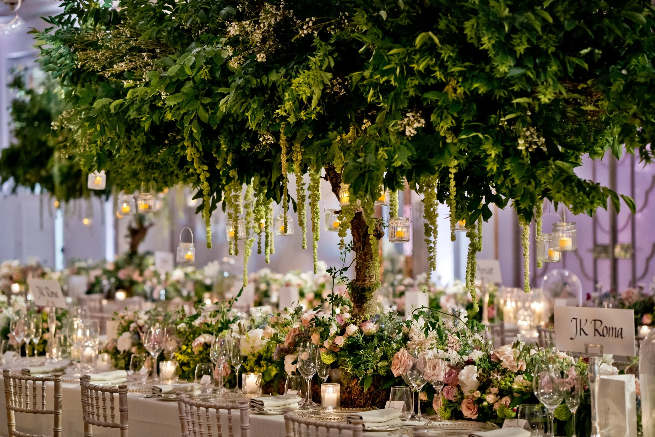 Elegant indoor wedding reception decorated with a large floral centerpiece resembling a small tree, with hanging lanterns and candles, surrounded by tables adorned with flowers, glassware, and white chairs.