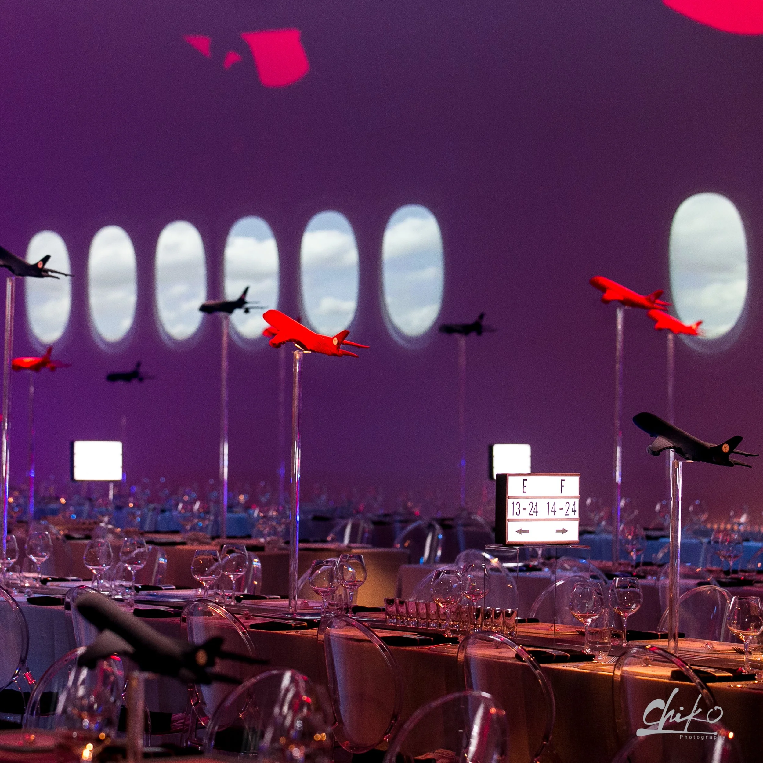 Multiple transparent chairs arranged in rows at an airport terminal or waiting area with large windows in the background, and flight information sign and airplane models hanging from the ceiling.