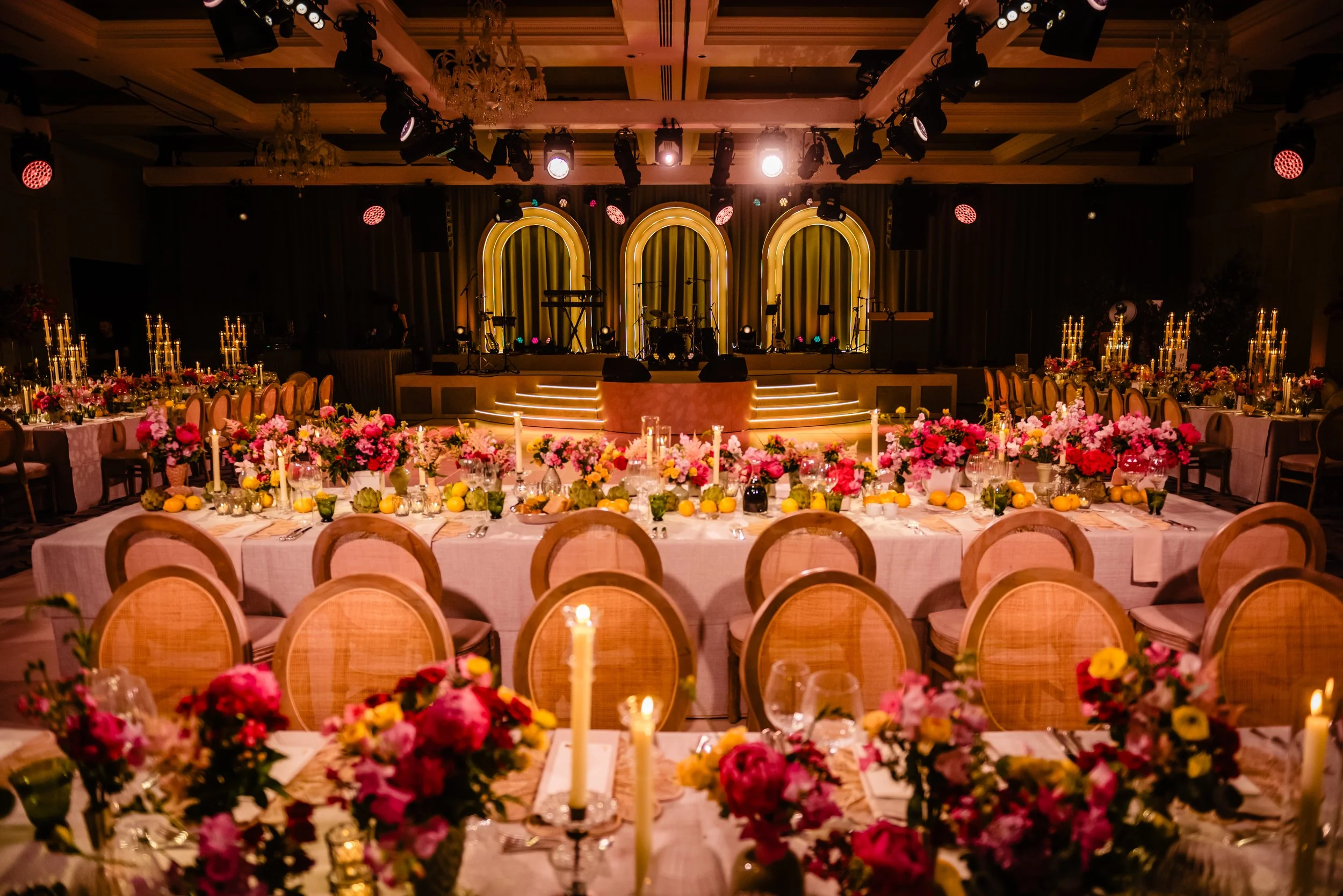 Elegant banquet hall decorated with flowers, candles, and lighting, with a stage set for a live performance.