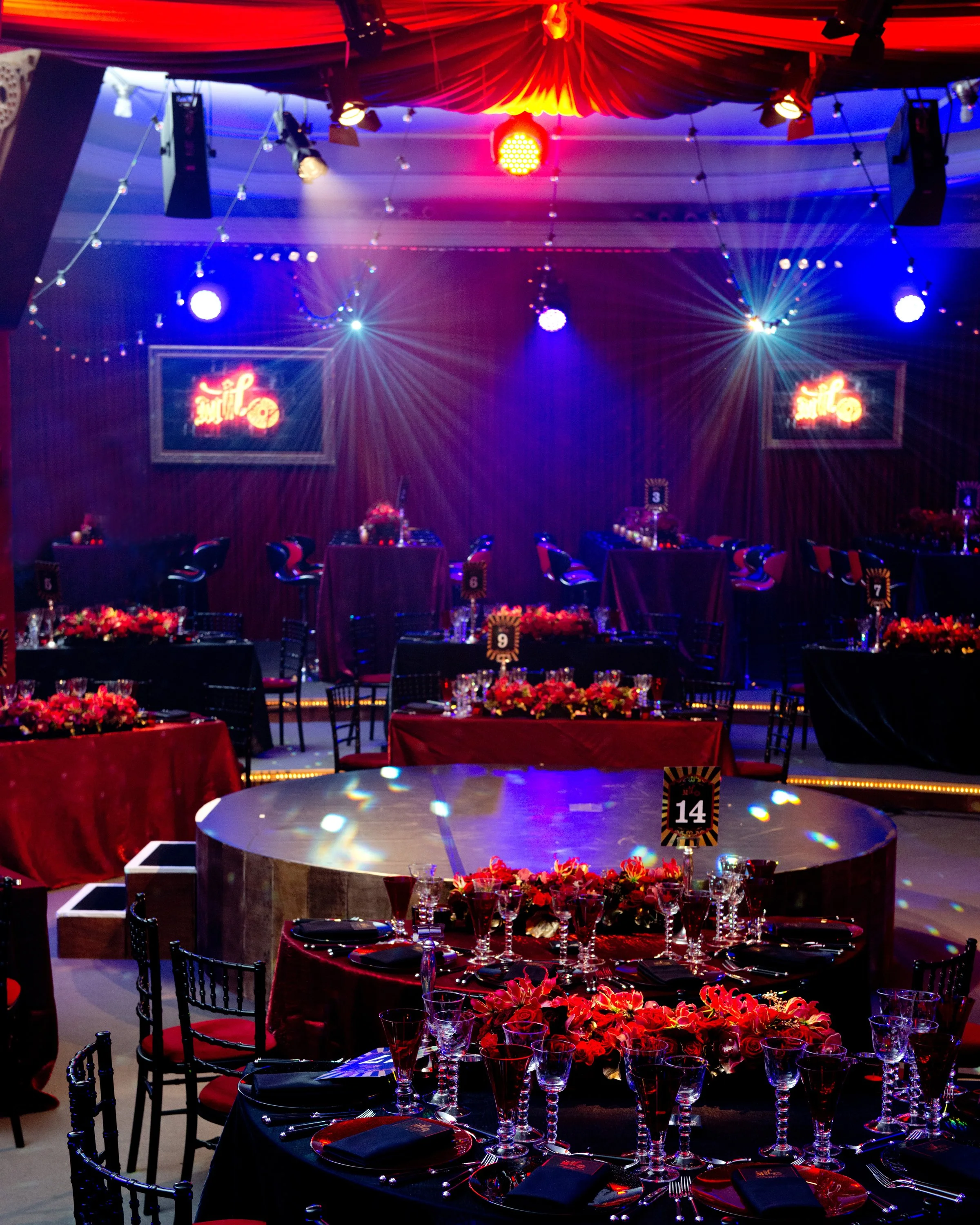 A decorated event space with round tables covered in red tablecloths, set with elaborate glassware, black napkins, and floral centerpieces featuring red flowers. The background features a stage with colorful lighting, including blue and red spotlight