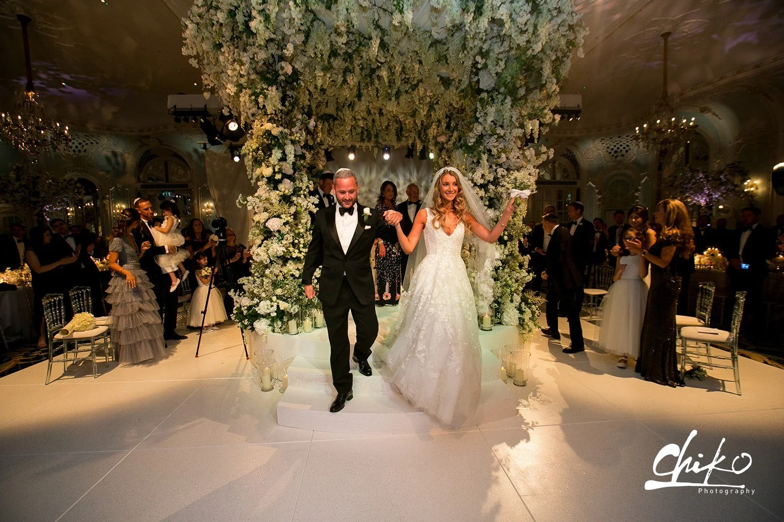 A newlywed couple walking down the aisle at their wedding reception, holding hands and smiling. The bride is wearing a white lace gown with a veil; the groom is in a black tuxedo. The decorated arch is covered in white flowers and greenery, surrounde