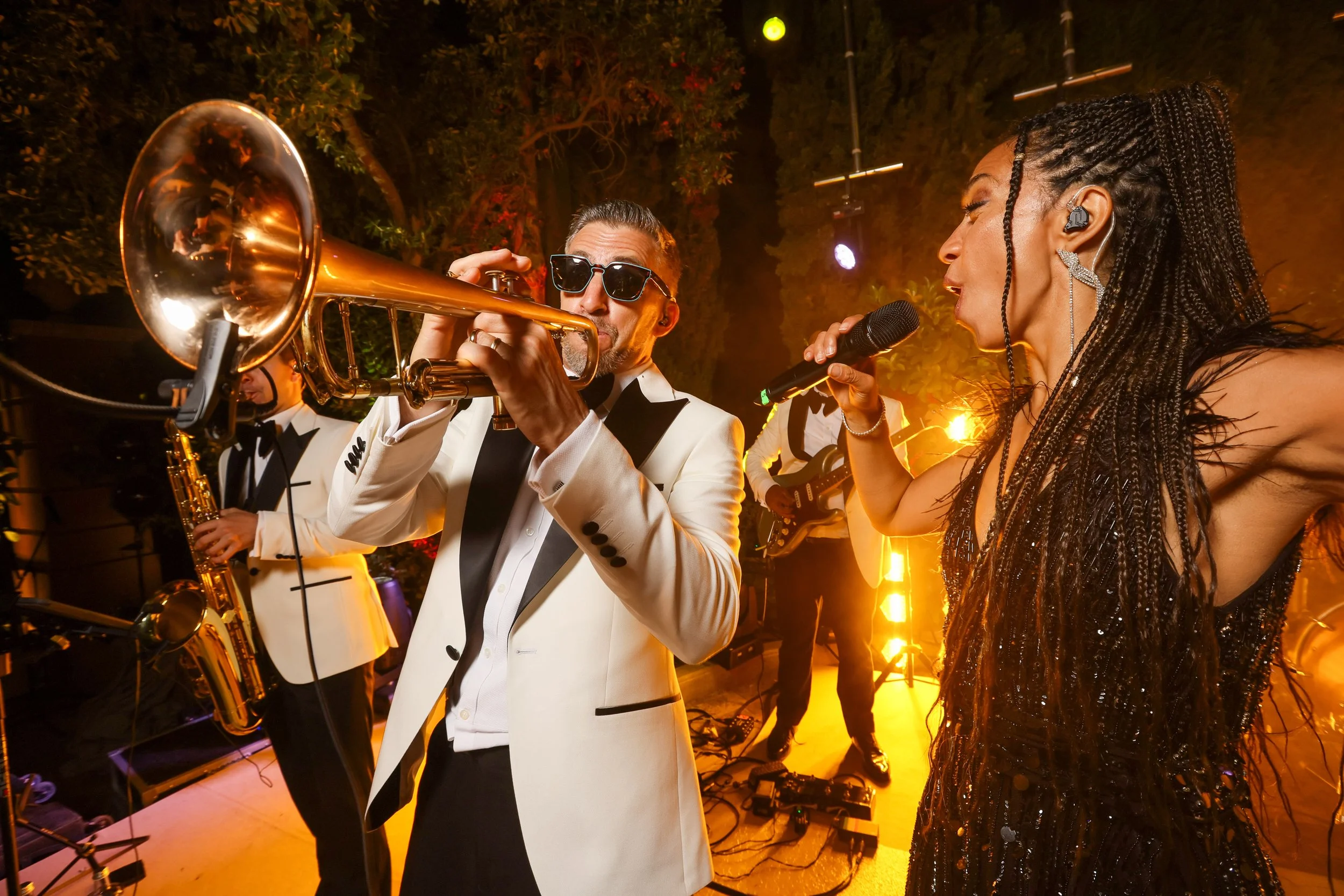 A man in a tuxedo playing a trumpet and a woman in a black dress singing into a microphone during a live music performance at a nighttime event.