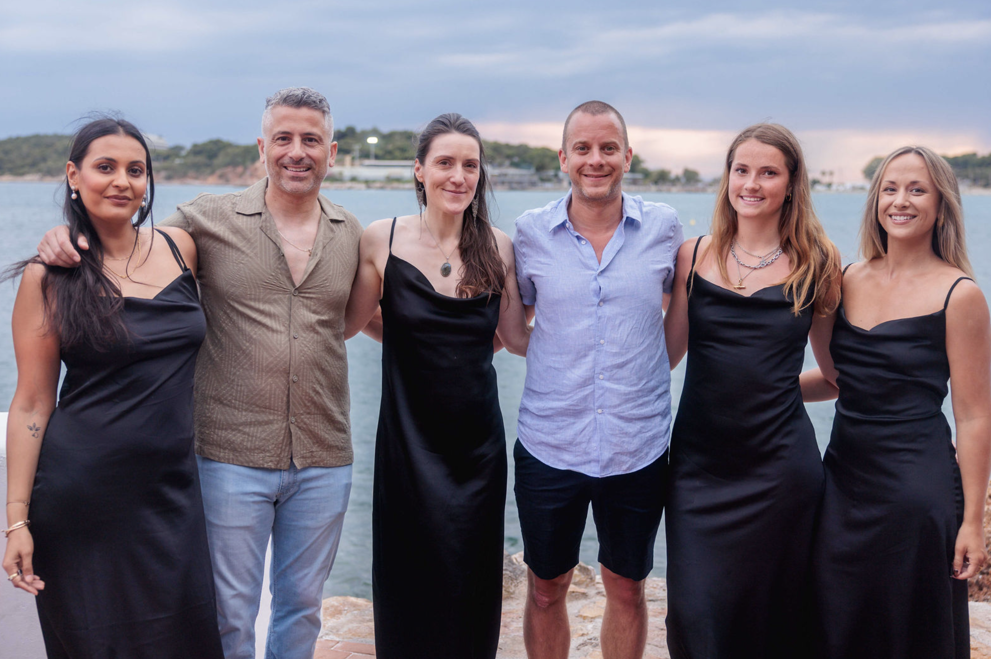 Group of six people standing outdoors near water, smiling for a photo with a scenic background of trees, water, and a cloudy sky.