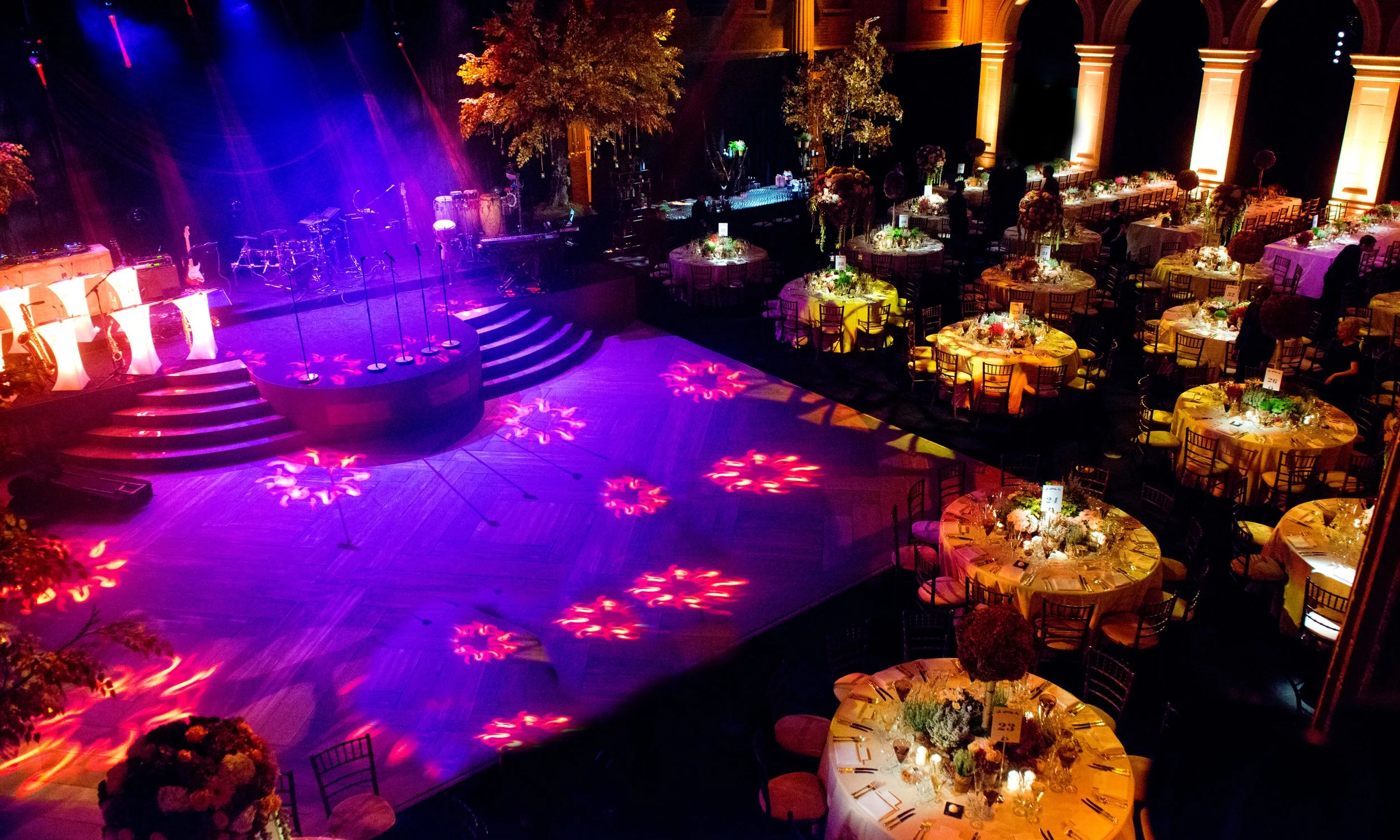 Elegant banquet hall set for a formal event with multiple round tables decorated with floral centerpieces, draped tablecloths, and candles. A stage with musical instruments and colorful lighting is visible in the background, with large trees and warm