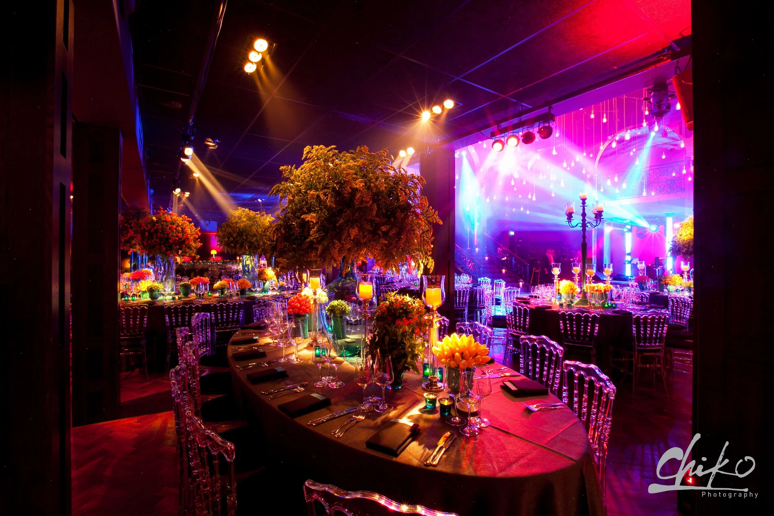 Elegant event venue decorated with colorful flower arrangements on round tables, ambient lighting, and stage lighting with purple and blue hues.