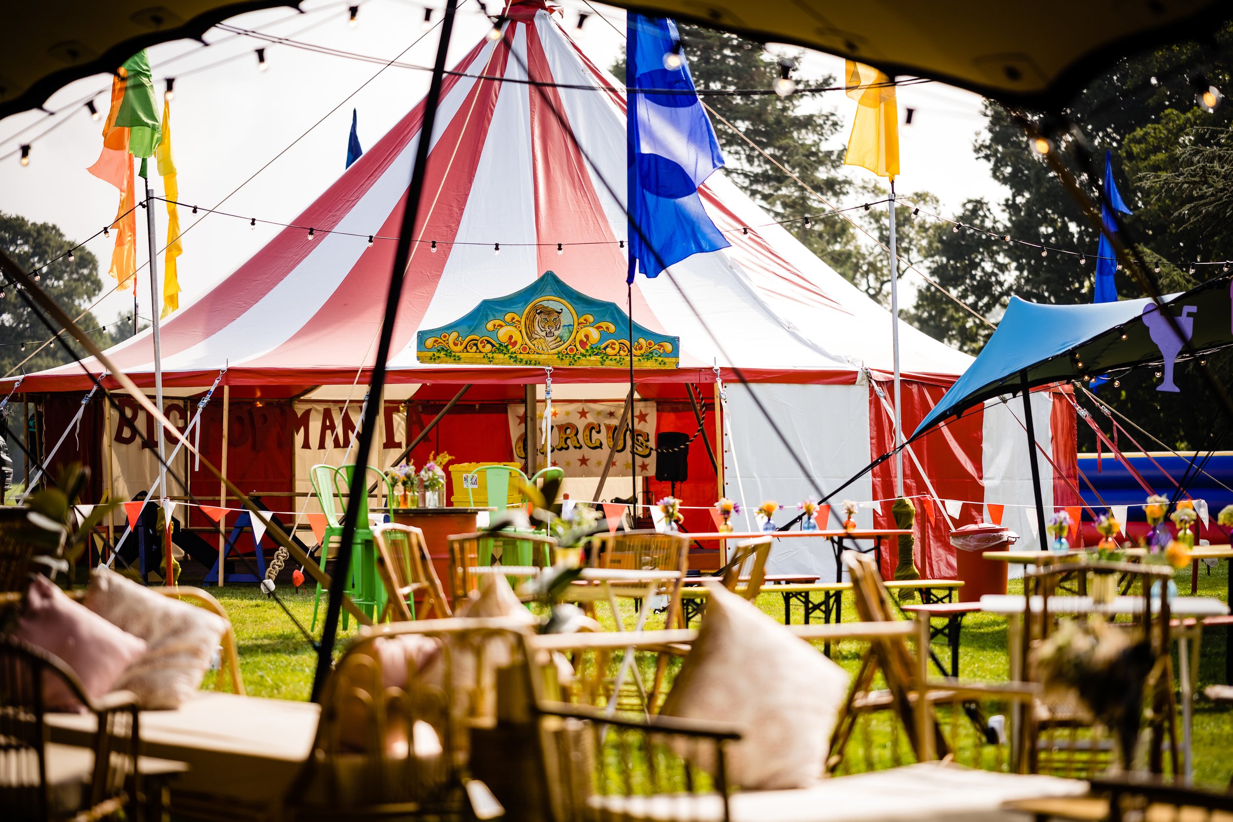 A colorful circus tent with red and white stripes, surrounded by yellow, blue, orange, and green flags, and outdoor seating with table and chairs, set up for an event in a park-like area with trees in the background for a Bar Mitzvah at  Wrotham Park