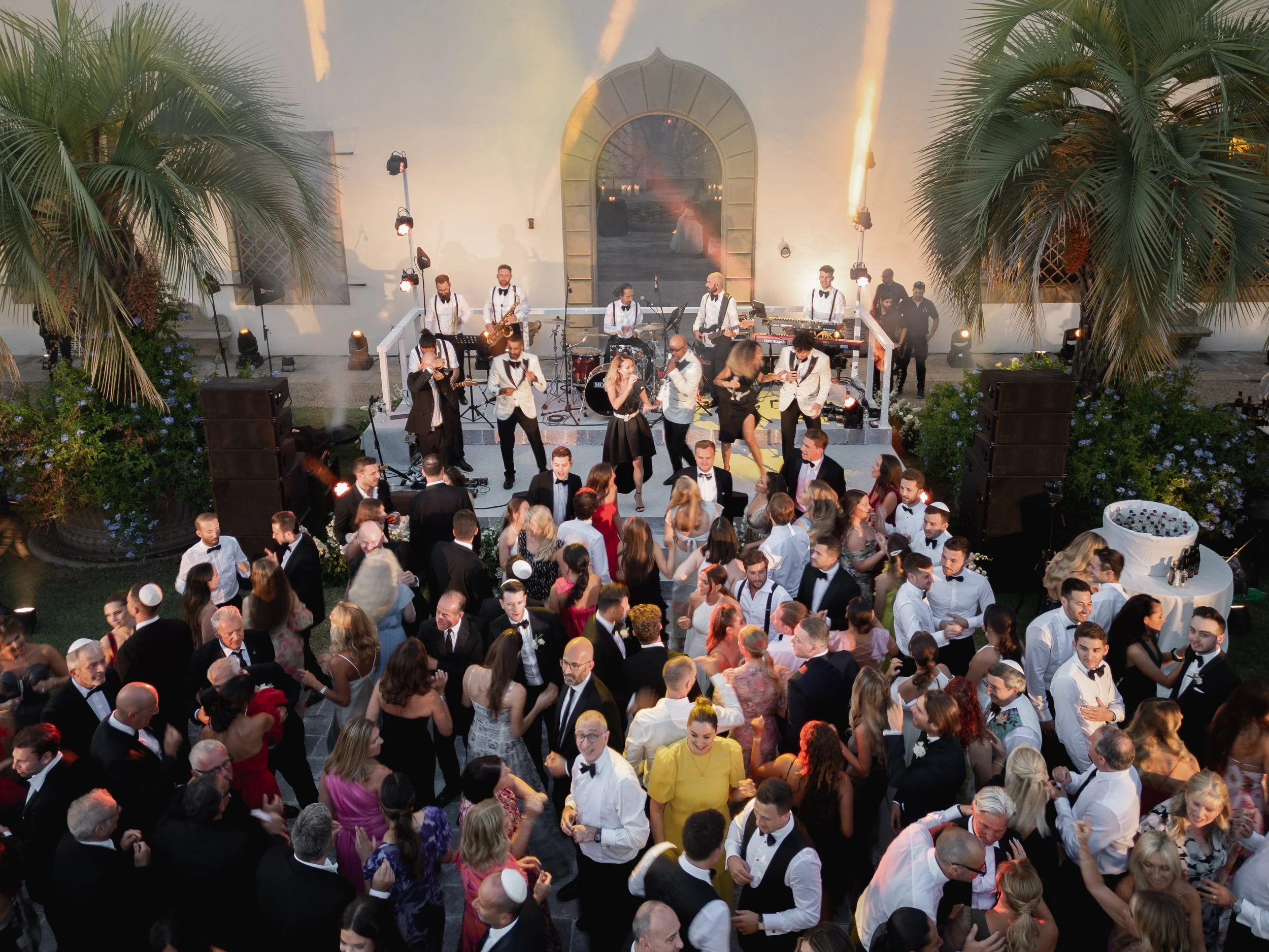 A large crowd of people dressed in formal attire dancing at an outdoor wedding reception with live band performing on stage and tropical plants and lighting creating a festive atmosphere.