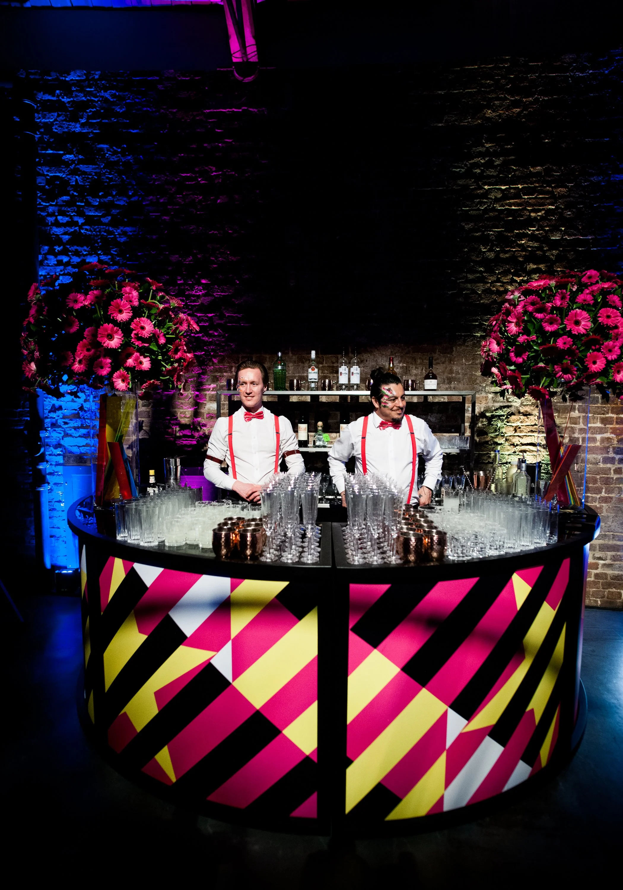 A colorful bar setup with two men dressed as bartenders, standing behind a vibrant pink, yellow, black, and white patterned bar counter. There are large pink flower arrangements on each side of the bar, with a brick wall background illuminated by pur