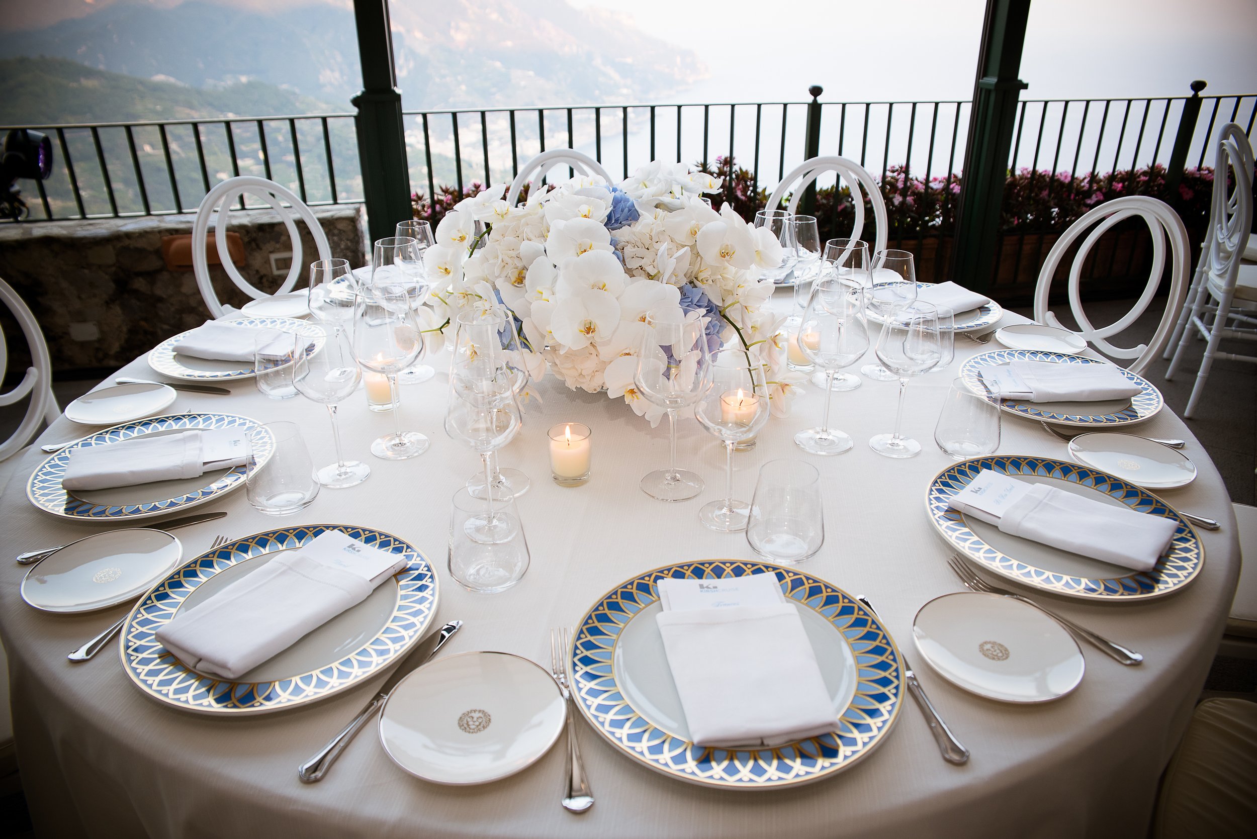 Elegantly set round dining table with white linens, gold and blue patterned china, white napkins, wine glasses, and small candles, overlooking a mountainous landscape through a balcony.