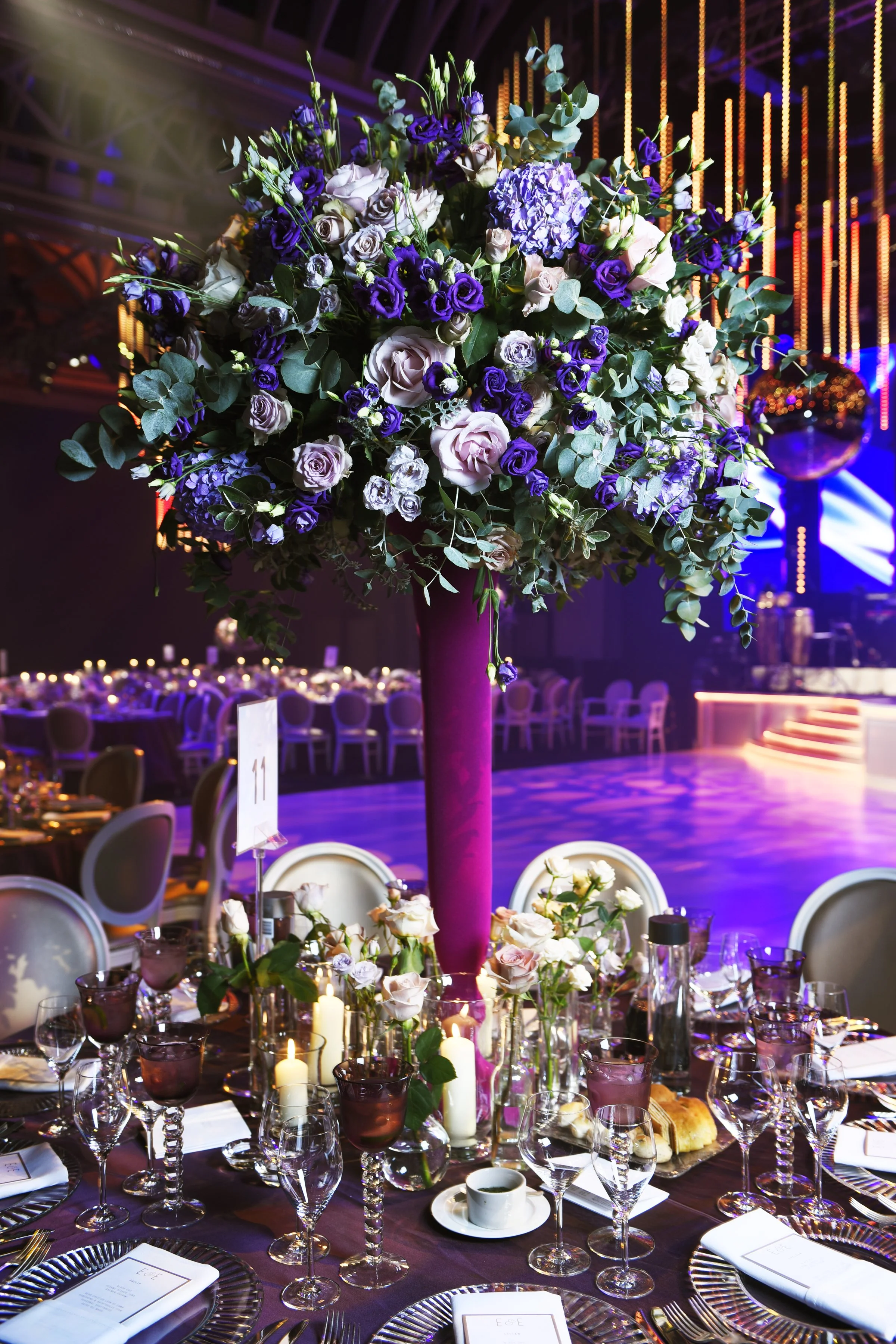 A large floral centerpiece with purple, white, and green flowers on a banquet table in a decorated event space with purple lighting and seating.