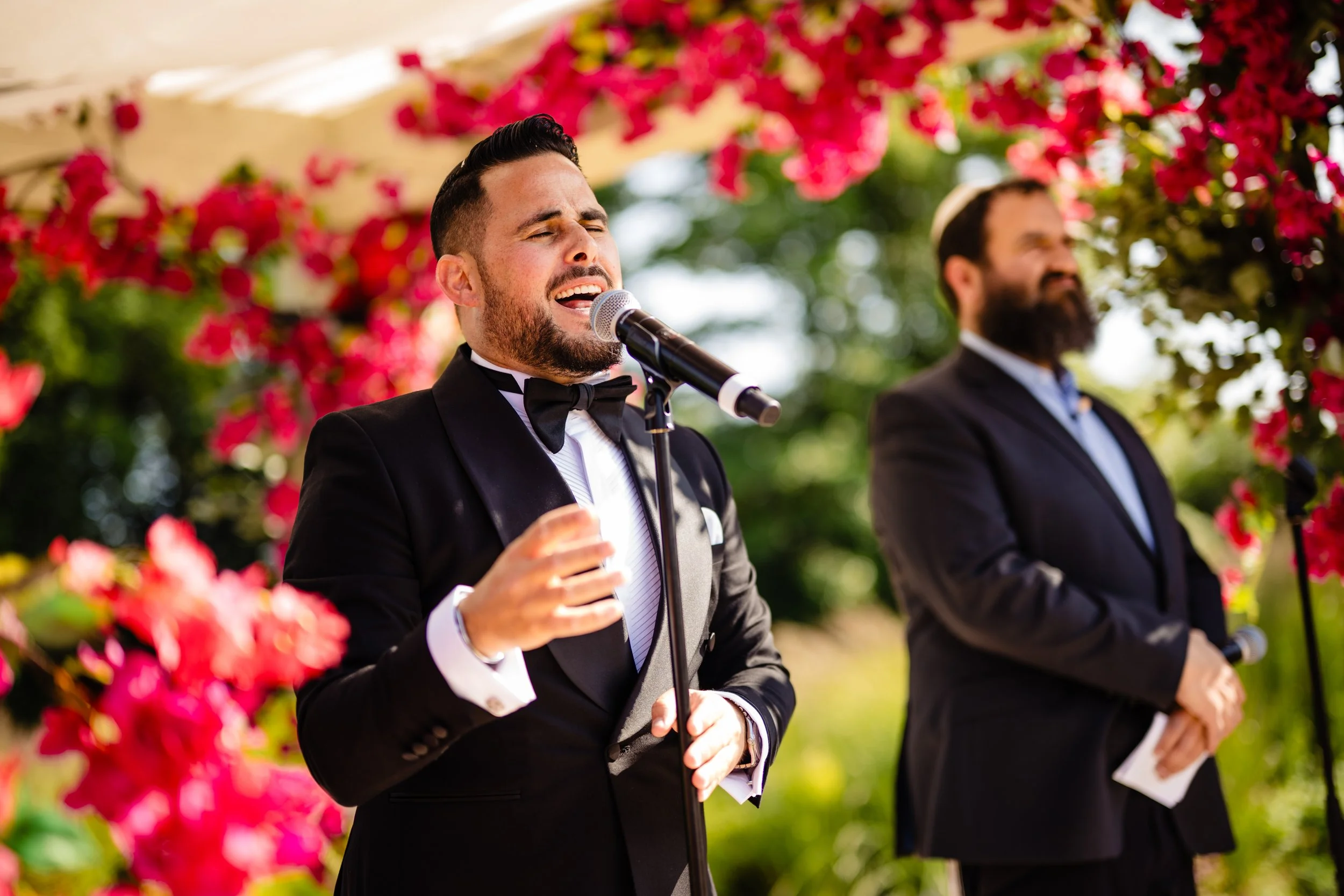 A man in a tuxedo singing into a microphone outdoors, with another man in a tuxedo standing nearby, surrounded by pink and red flowers and greenery Fairmont Windsor Park