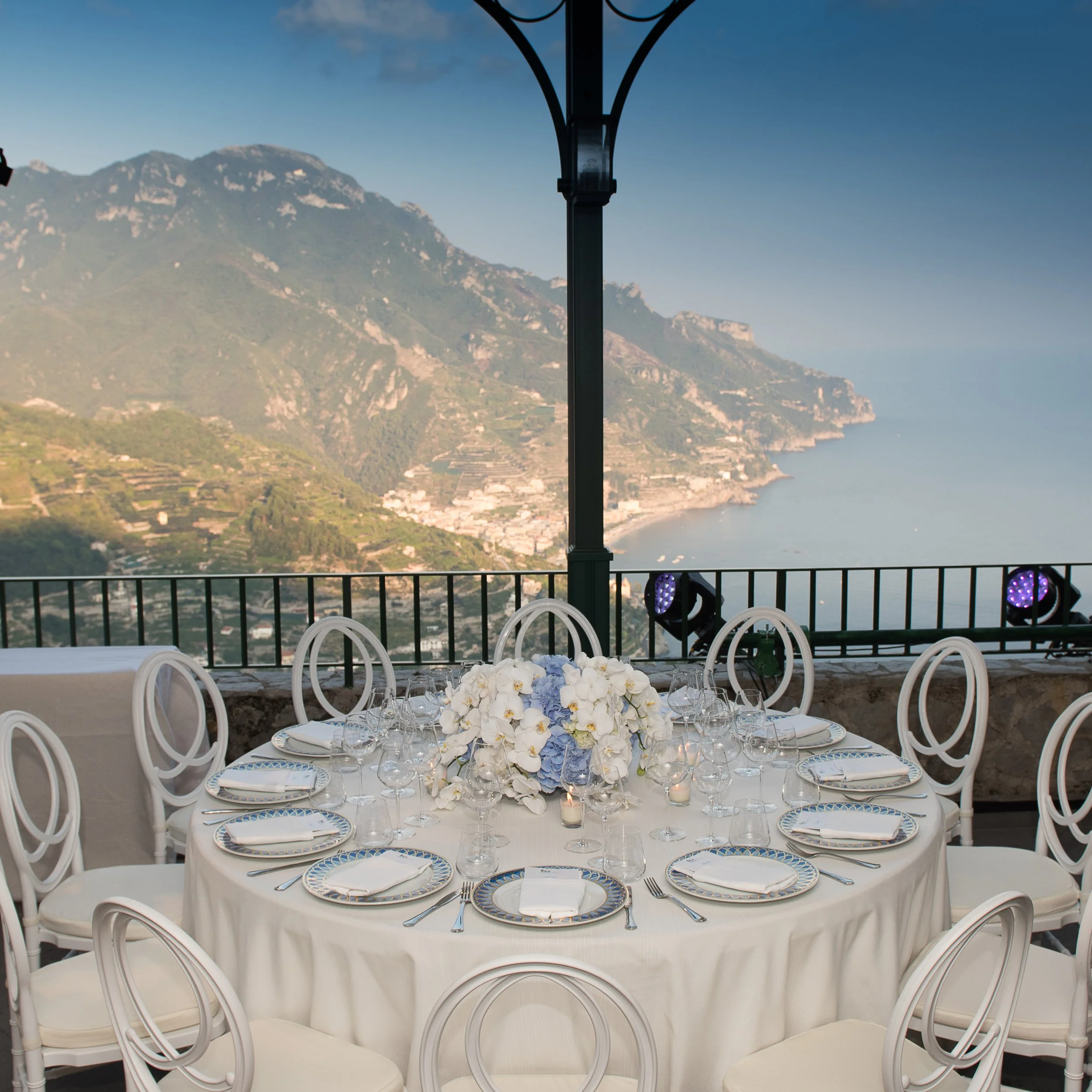 Elegant outdoor dining setup with a round table covered in a white tablecloth, adorned with a floral centerpiece of white and blue flowers, set with plates, wine glasses, and cutlery. White chairs surround the table, overlooking a scenic mountain lan