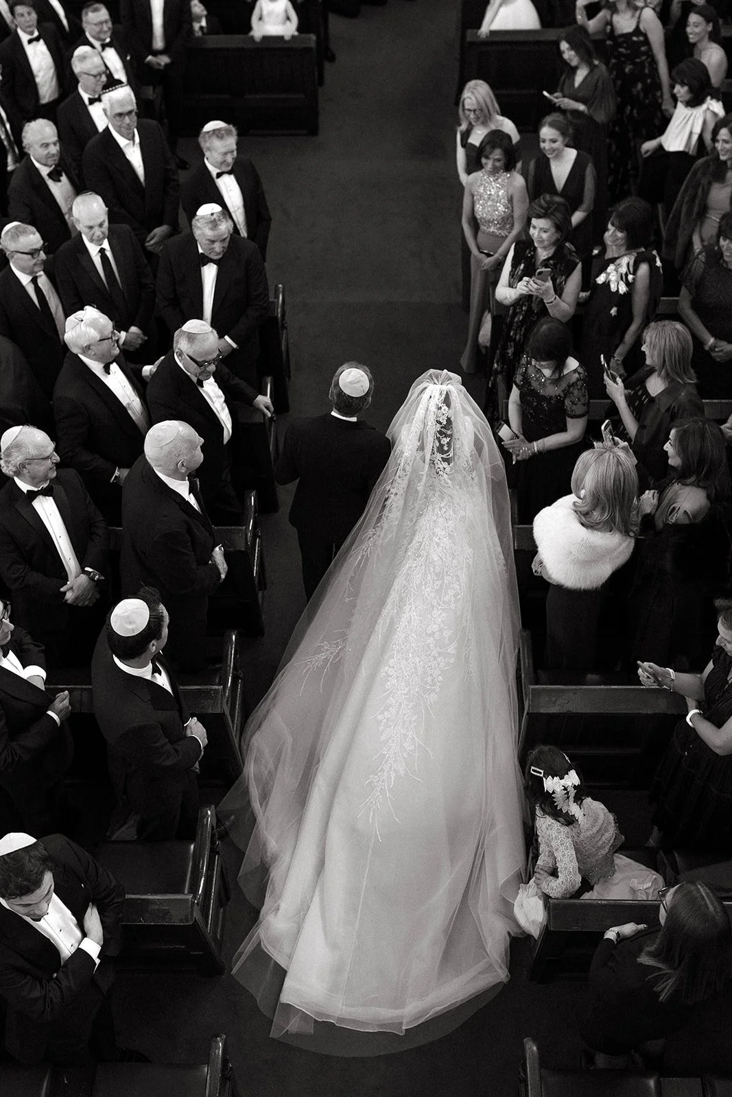 Black and white photo of a wedding ceremony in a church, with a bride in a long wedding gown and veil walking down the aisle with a man, surrounded by guests in formal attire, some taking photos  for the recpetion to be at The Peninsula London
