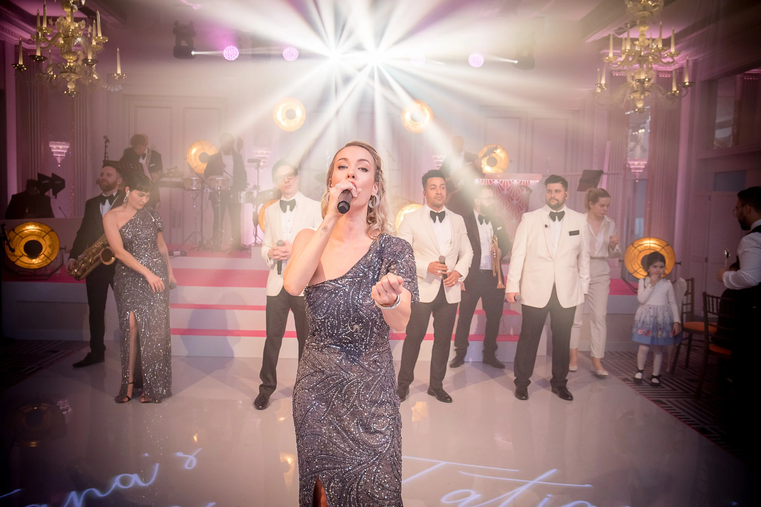A woman in a sparkly evening gown singing into a microphone on a stage with a band and dancers, set in an elegant room with chandeliers and lighting effects.