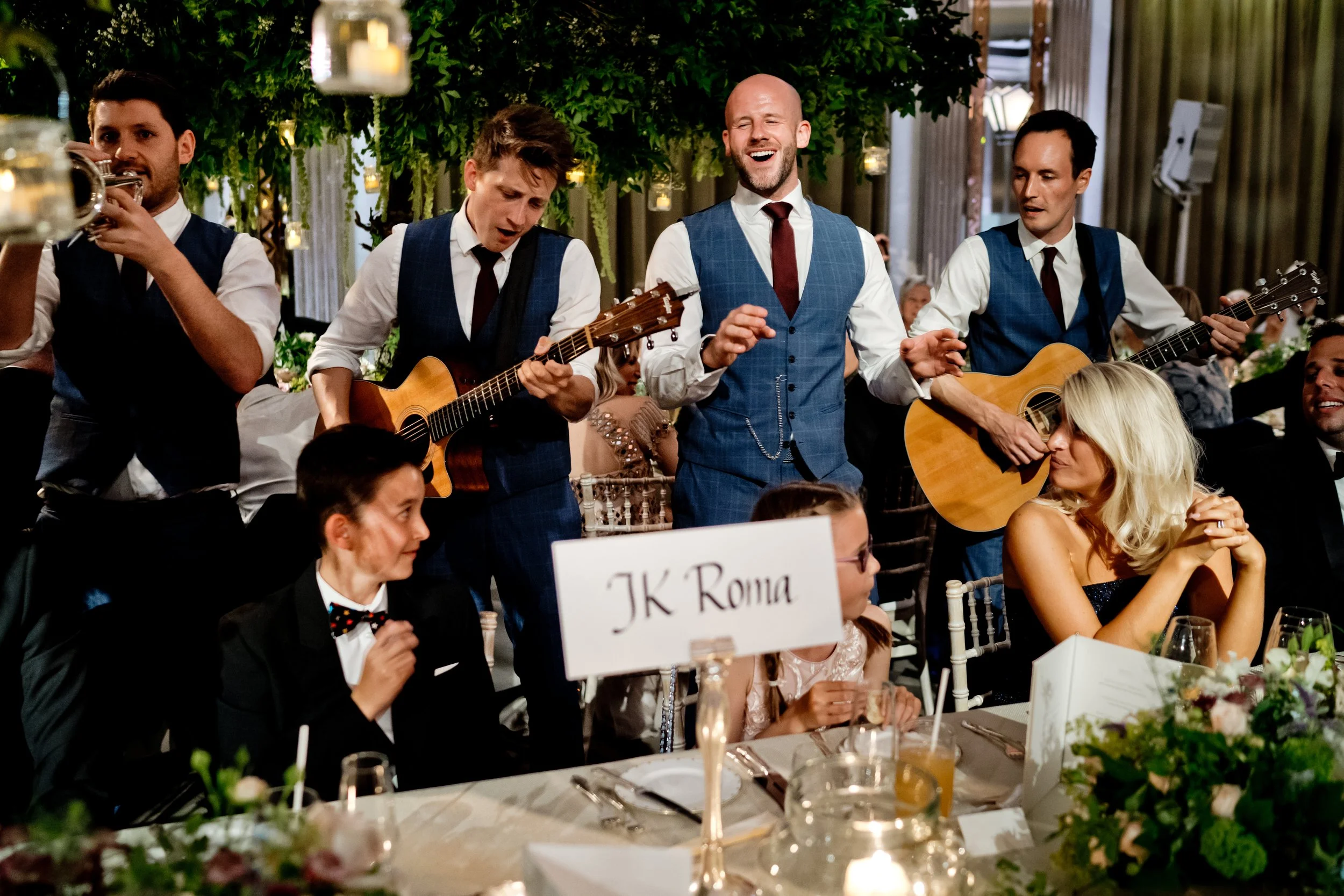 A group of musicians playing guitars and singing at a wedding reception, with guests seated at tables decorated with floral centerpieces and candles.