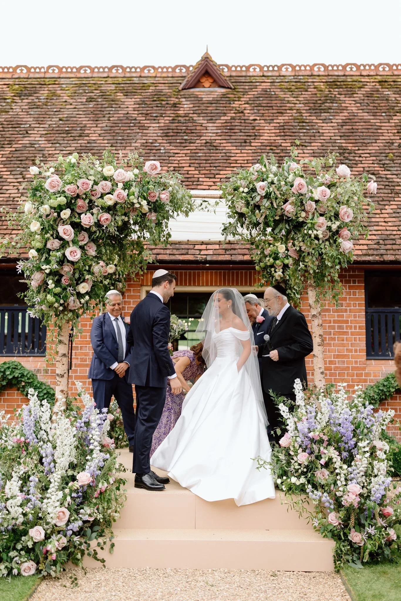 A wedding ceremony outdoors under a floral arch with pink and white roses, greenery, and flowering plants, with a bride and groom, officiant, and guests present.