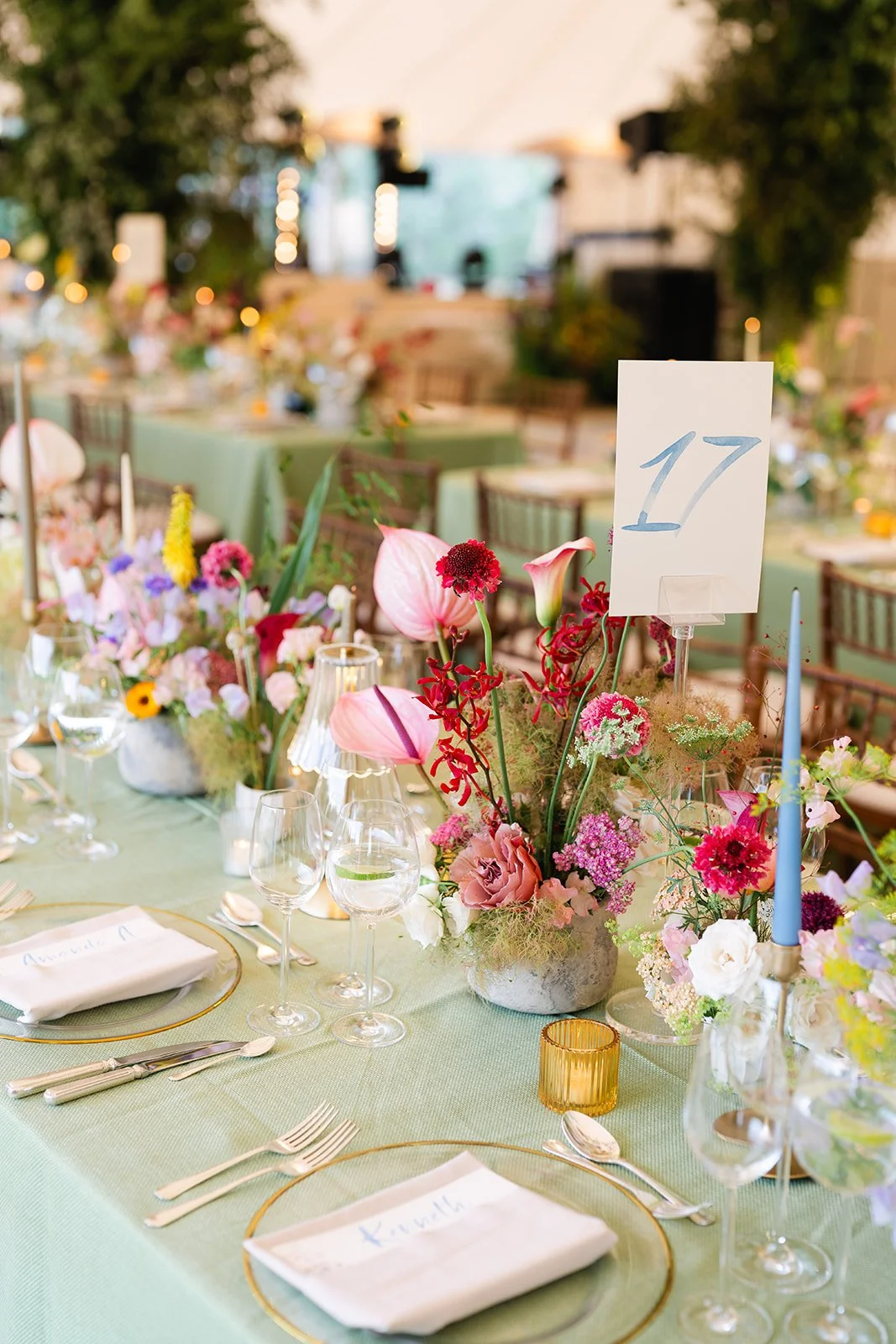 Elegant banquet table decorated with pink, purple, and red flowers, set with glassware, silverware, and cloth napkins, with a table number card displaying '17', in a softly lit, floral-themed event space.