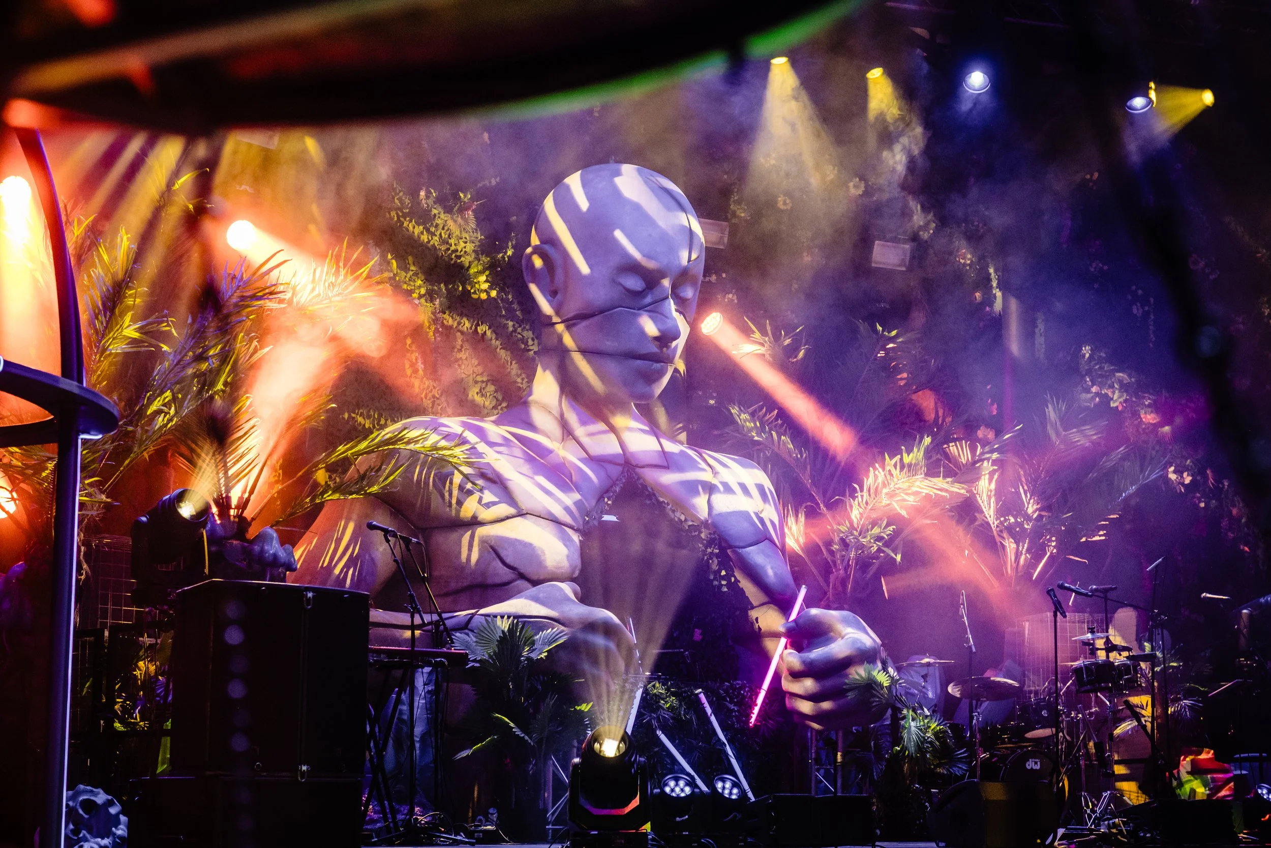 A large sculpture of a seated human face with closed eyes, illuminated by colorful stage lights, surrounded by greenery and stage equipment, for a luxury Bar Mitzvah 