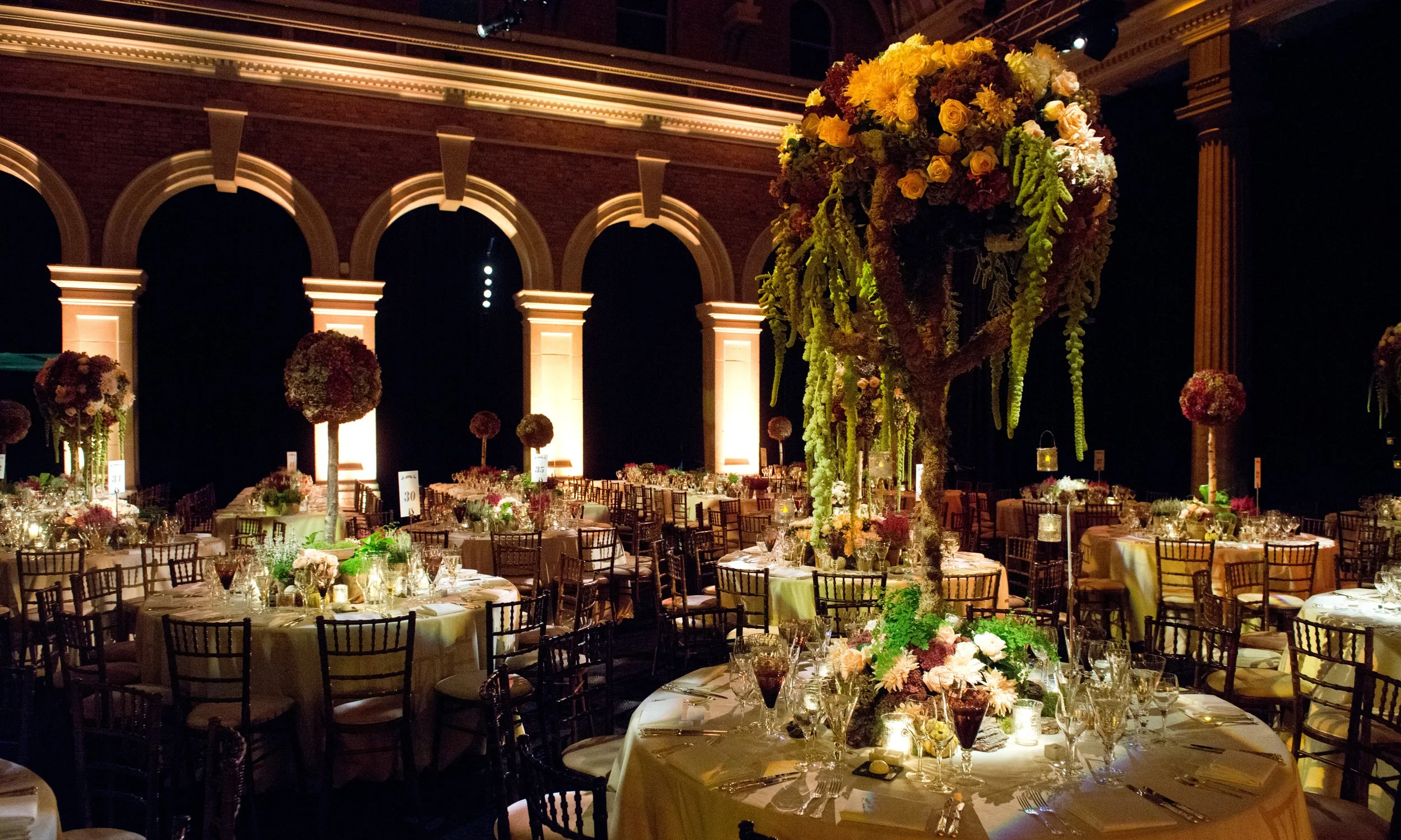 An elegantly decorated banquet hall with round tables covered in white tablecloths, adorned with floral centerpieces. There are numerous chairs arranged around the tables and a prominent tall floral arrangement in the center of the image, featuring y