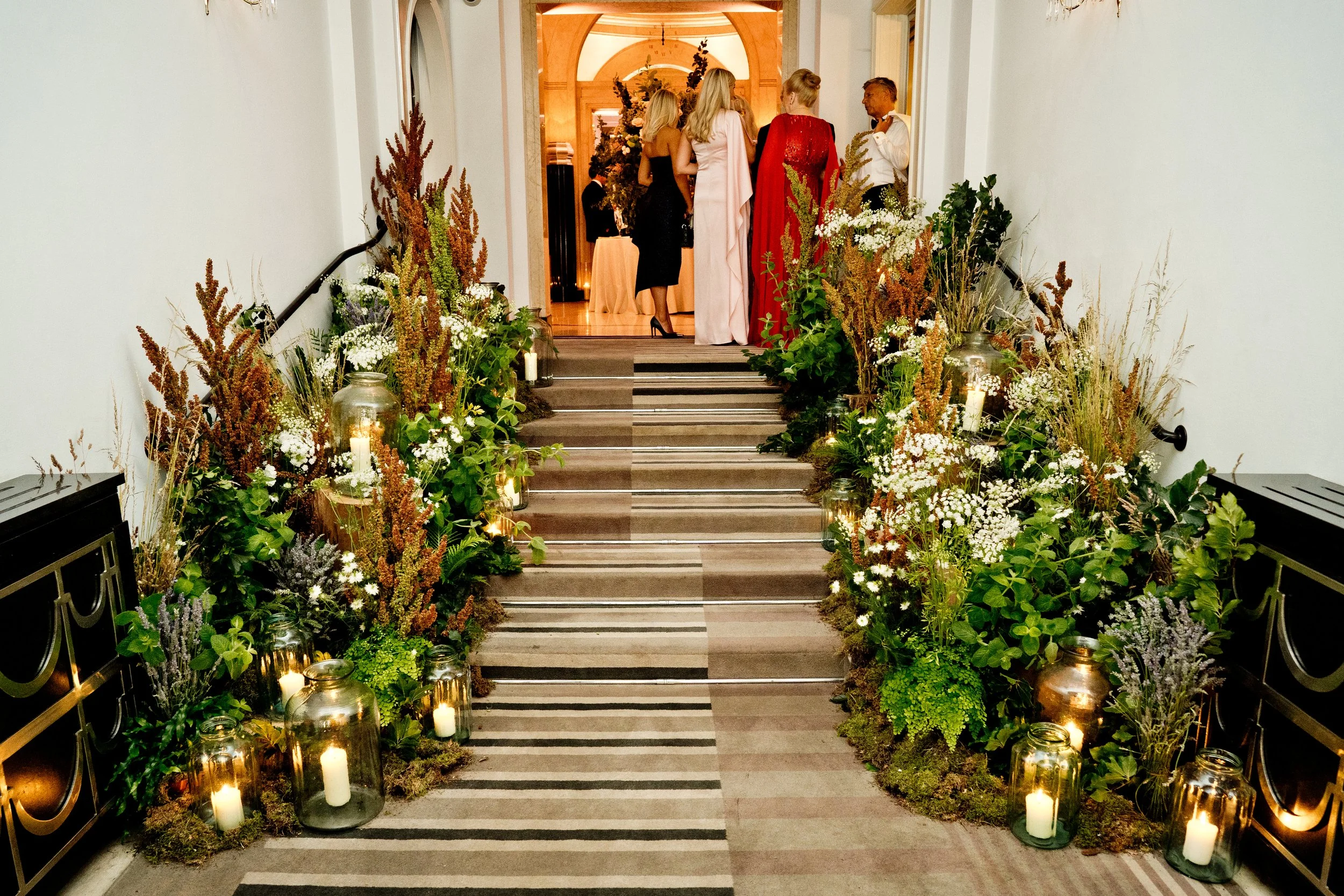A decorated indoor hallway with stairs leading up to a room, featuring numerous candles and plants along the sides, and a group of people standing at the top, engaged in conversation.