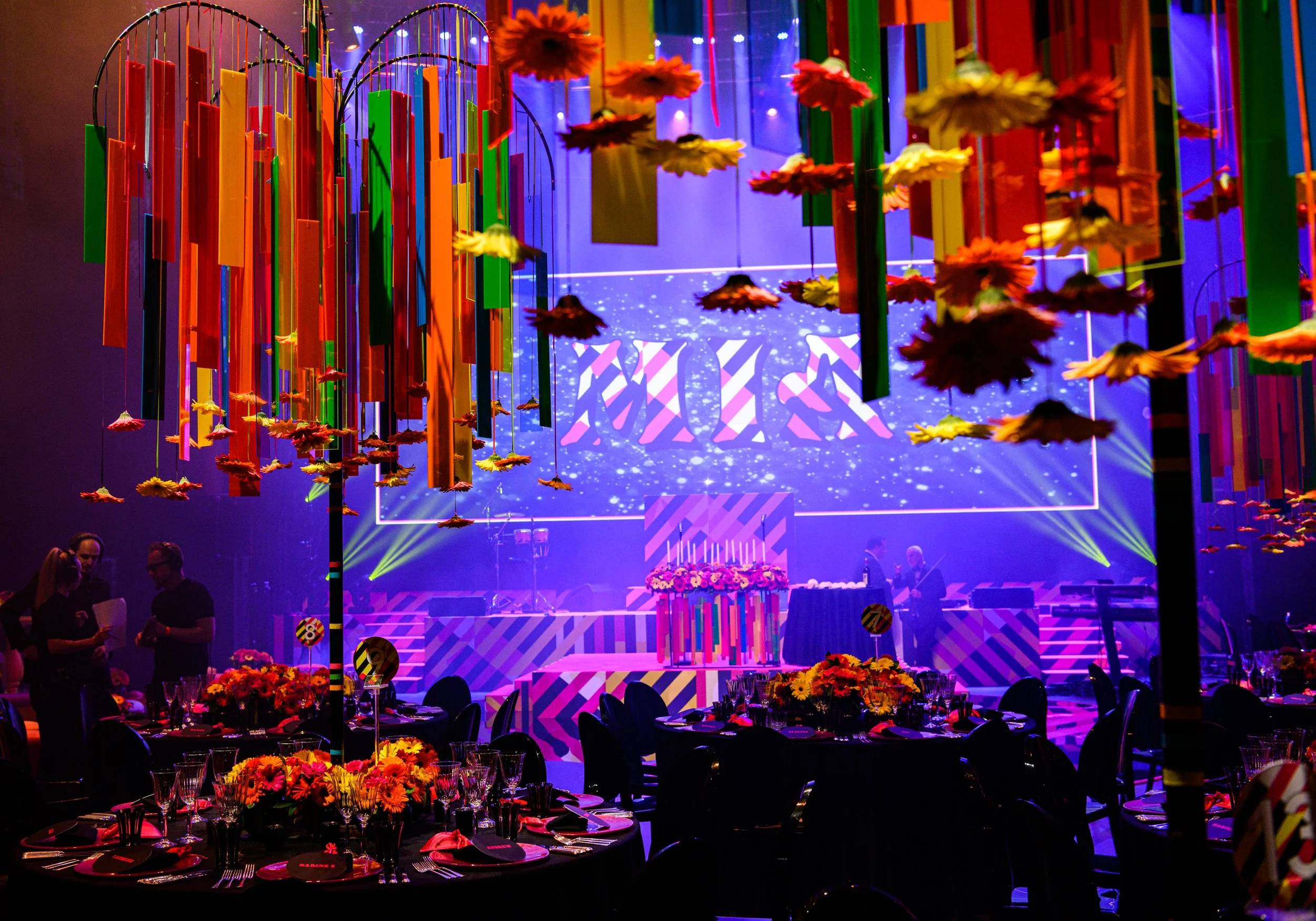 A brightly lit event space with colorful hanging decorations that resemble flowers and rectangular strips. There are round tables set with black tablecloths, plates, glasses, and floral centerpieces. In the background, a stage with a large illuminated screen displaying a colorful design is visible, along with a few people standing and talking.