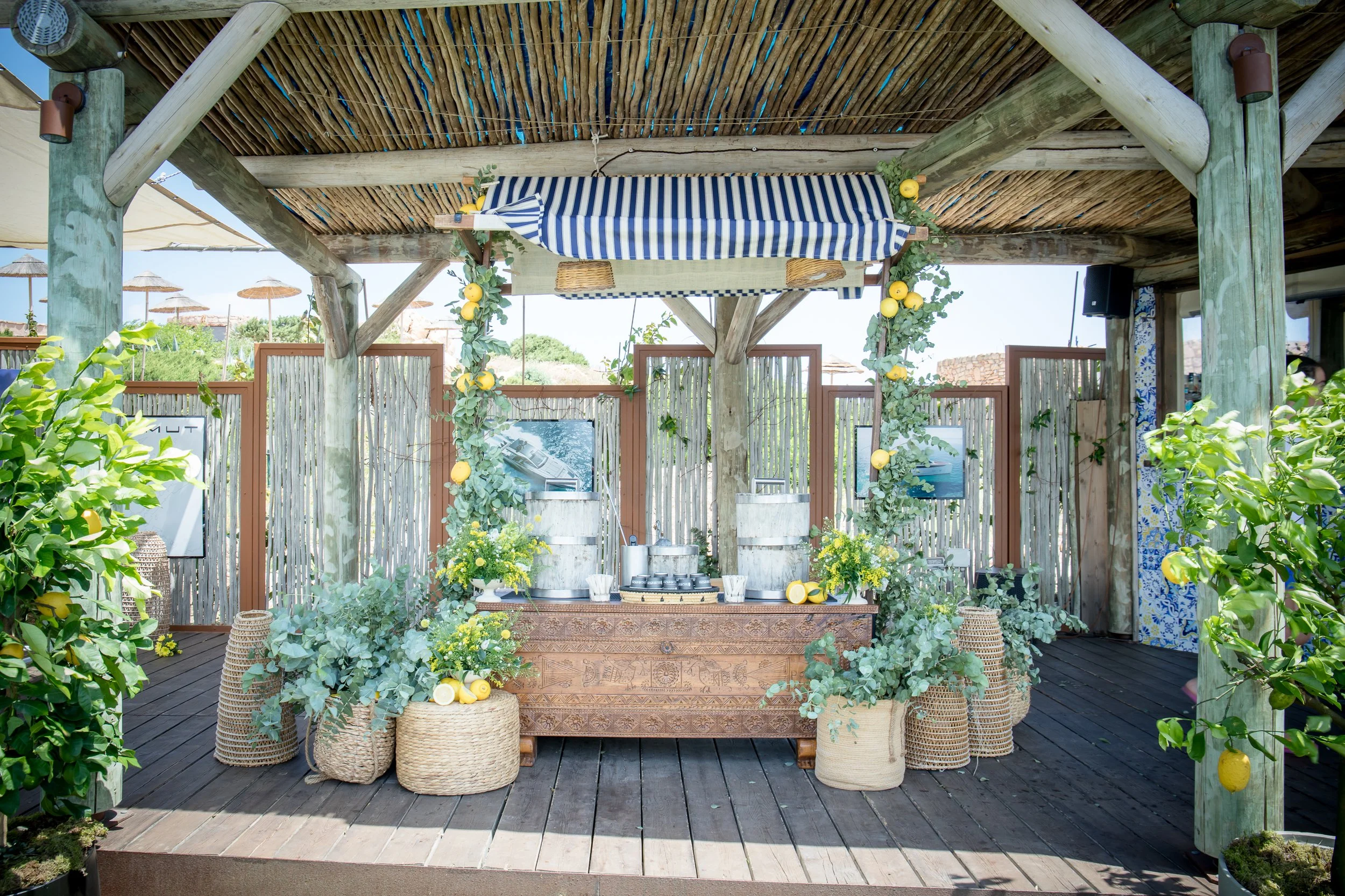 An outdoor bar or refreshment area with a wooden counter decorated with green foliage and yellow lemons, under a wooden canopy with rustic support beams. There are various wicker baskets and potted plants around, with a background of wooden and bambo