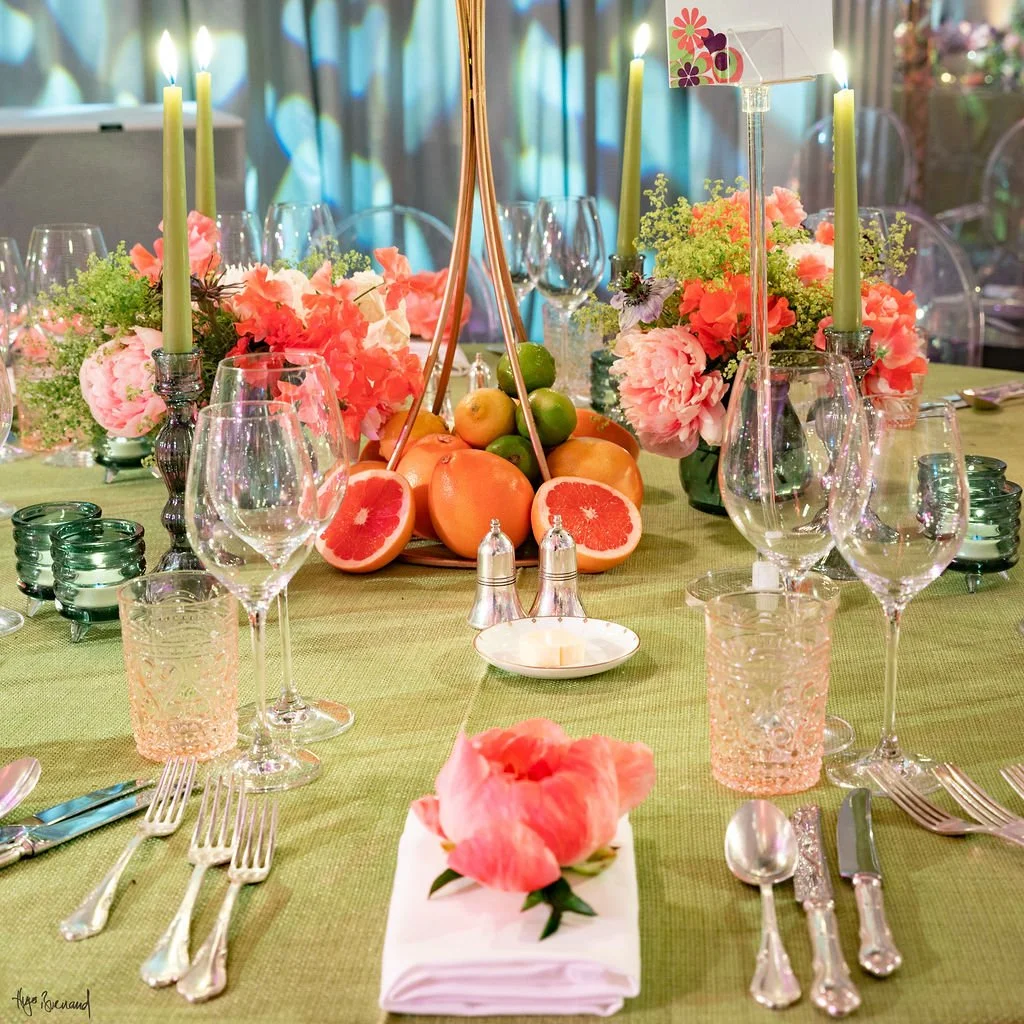 A decorated dining table set for a celebration with pink and green floral arrangements, tall green candles, a basket of citrus fruits including grapefruits, apples, and oranges, surrounded by wine glasses, water glasses, napkins with pink flowers, an