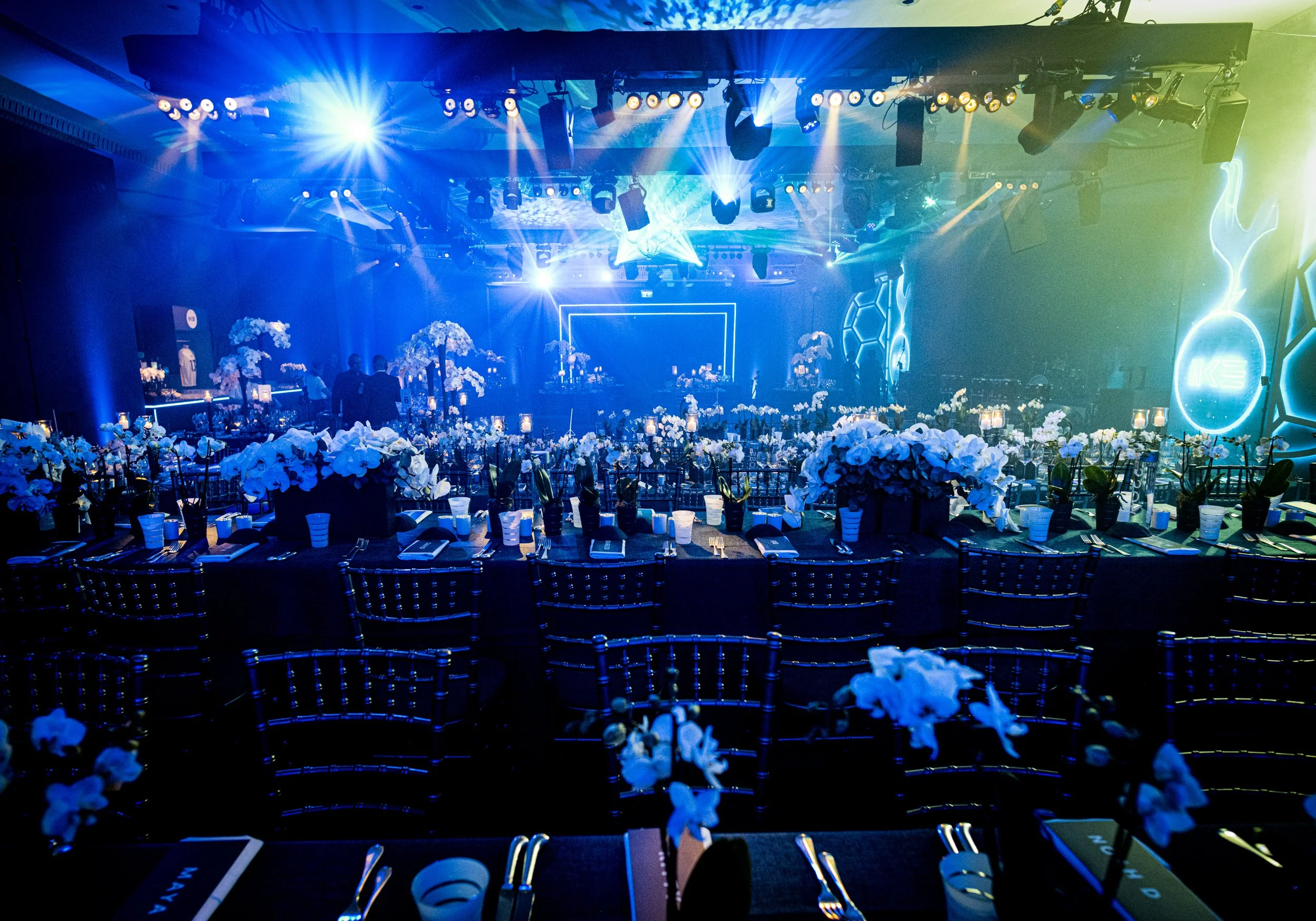 A large event space or banquet hall decorated for a celebration, featuring tables with white floral centerpieces, chairs, and blue lighting with spotlights and laser effects on the ceiling.