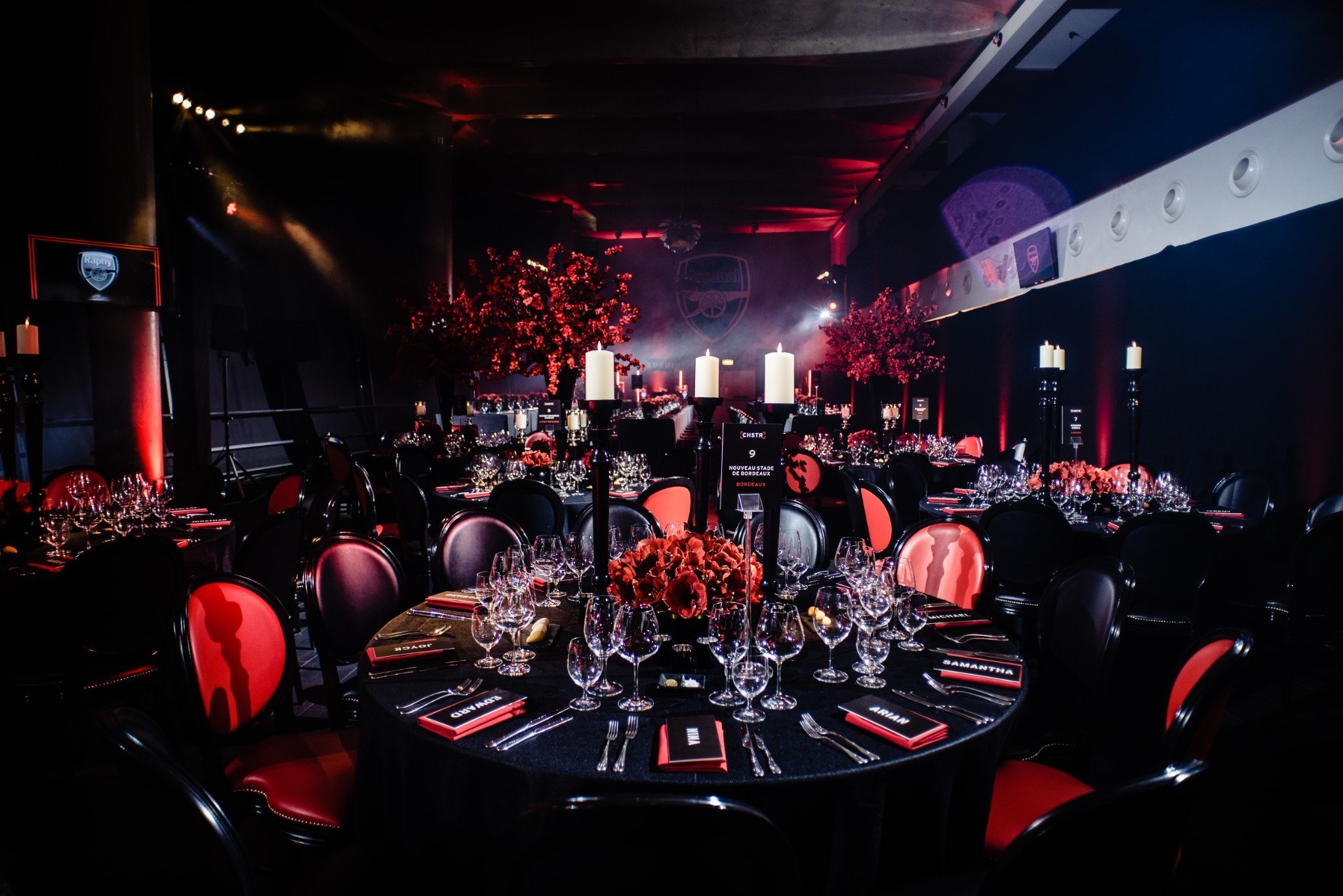 A darkly lit event space decorated with red and black theme, featuring tables set with wine glasses, candles, and floral arrangements, with a background displaying a logo of Arsenal Football Club.