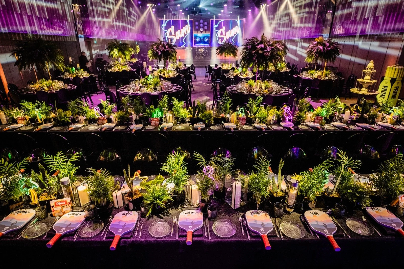An event space decorated for a celebration with tables adorned with greenery, neon signs, and flamingo decorations, with a stage in the background featuring two large screens and purple lighting.