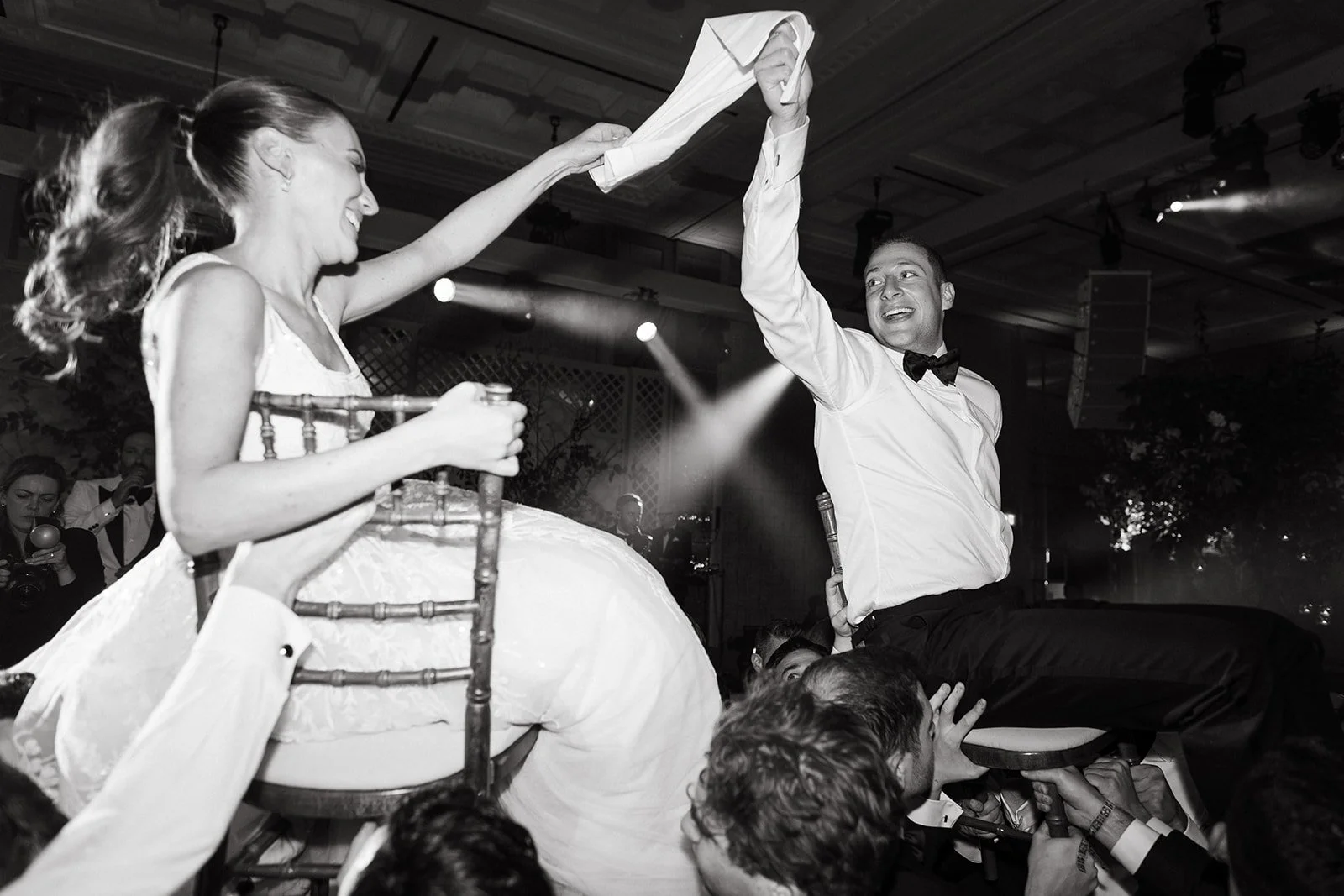 Bride and Groom in formal attire with a bow tie and is holding up the woman's arm, who is wearing a dress and seated on a chair. At The Peninsula London for the wedding reception with other people in the background