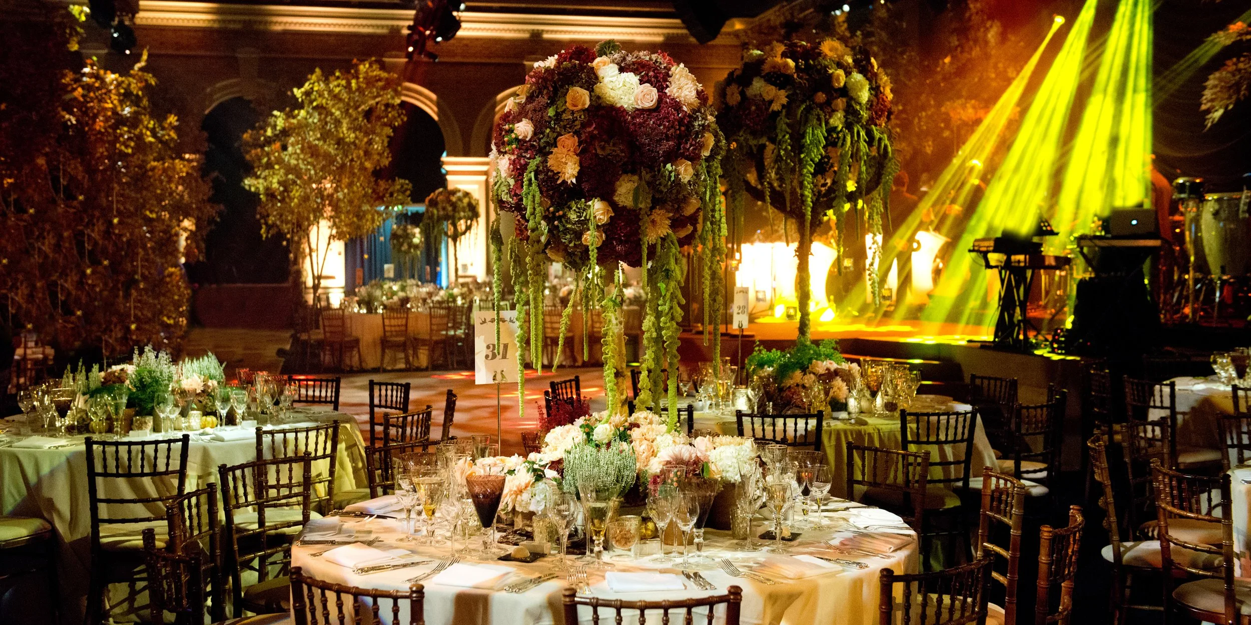 Elegant event hall with round tables decorated with floral centerpieces, surrounded by wooden chairs, and a large floral arrangement with hanging greenery at the center. Bright yellow stage lights are visible in the background.