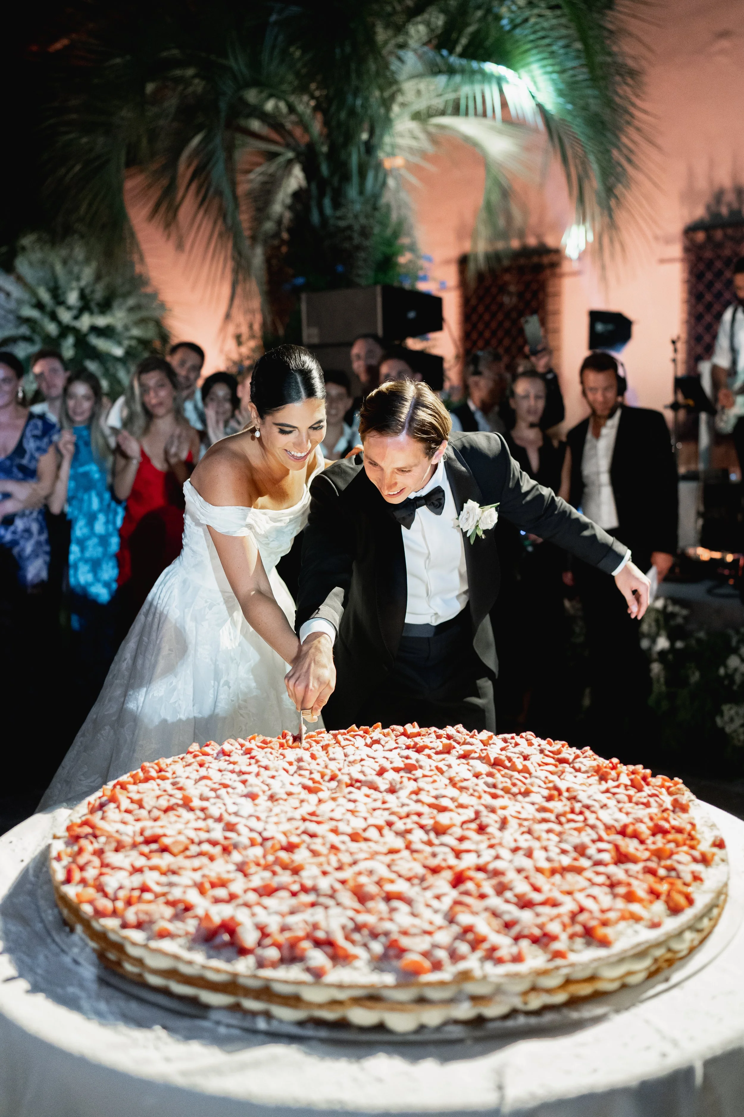A bride and groom cutting a large strawberry cheesecake surrounded by guests at a wedding reception at Villa Palmieri in Florence