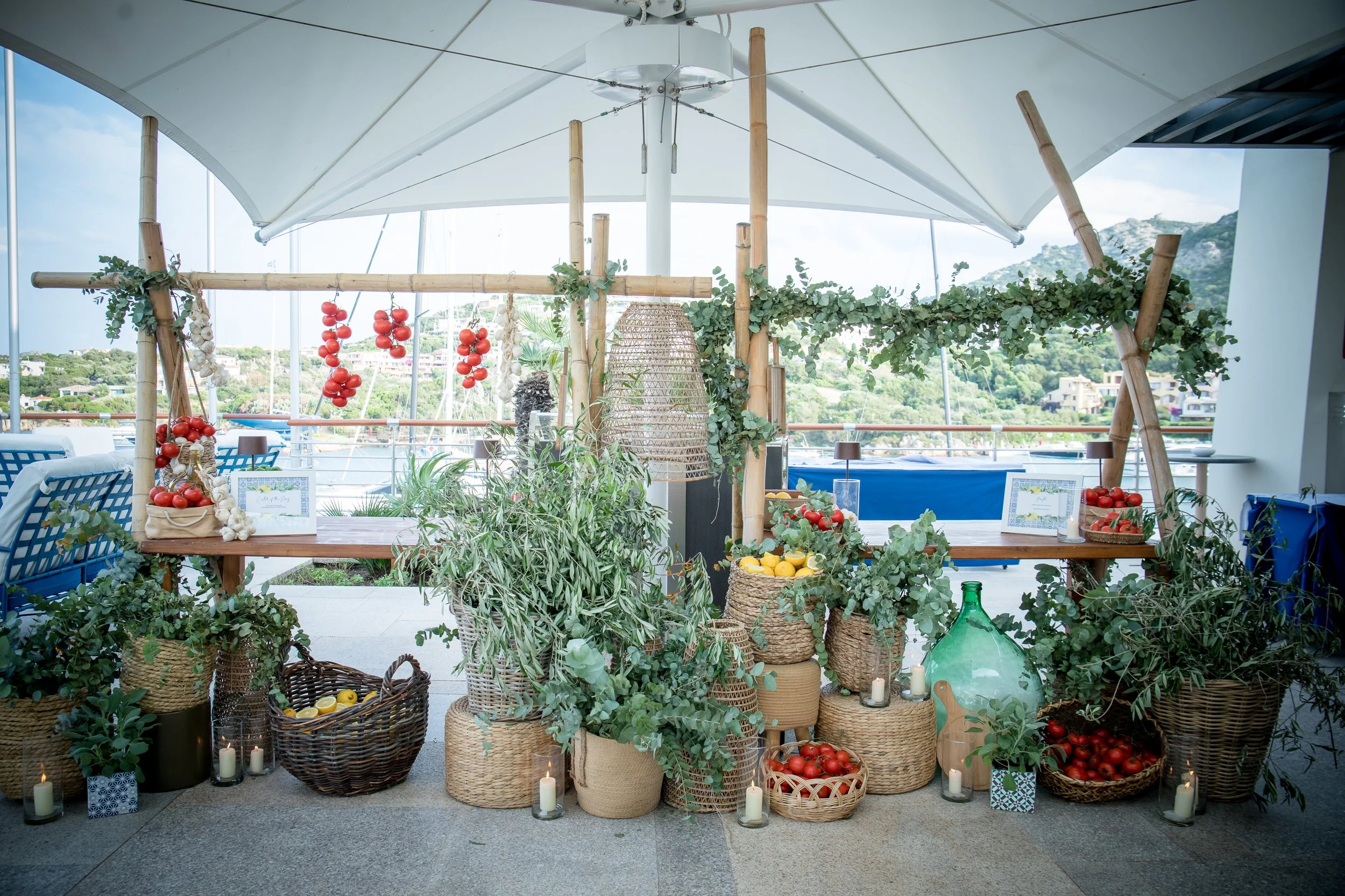 Display of fresh tomatoes, lemons, garlic, and greenery arranged in baskets and pots at an outdoor event with a scenic mountain view in the background.