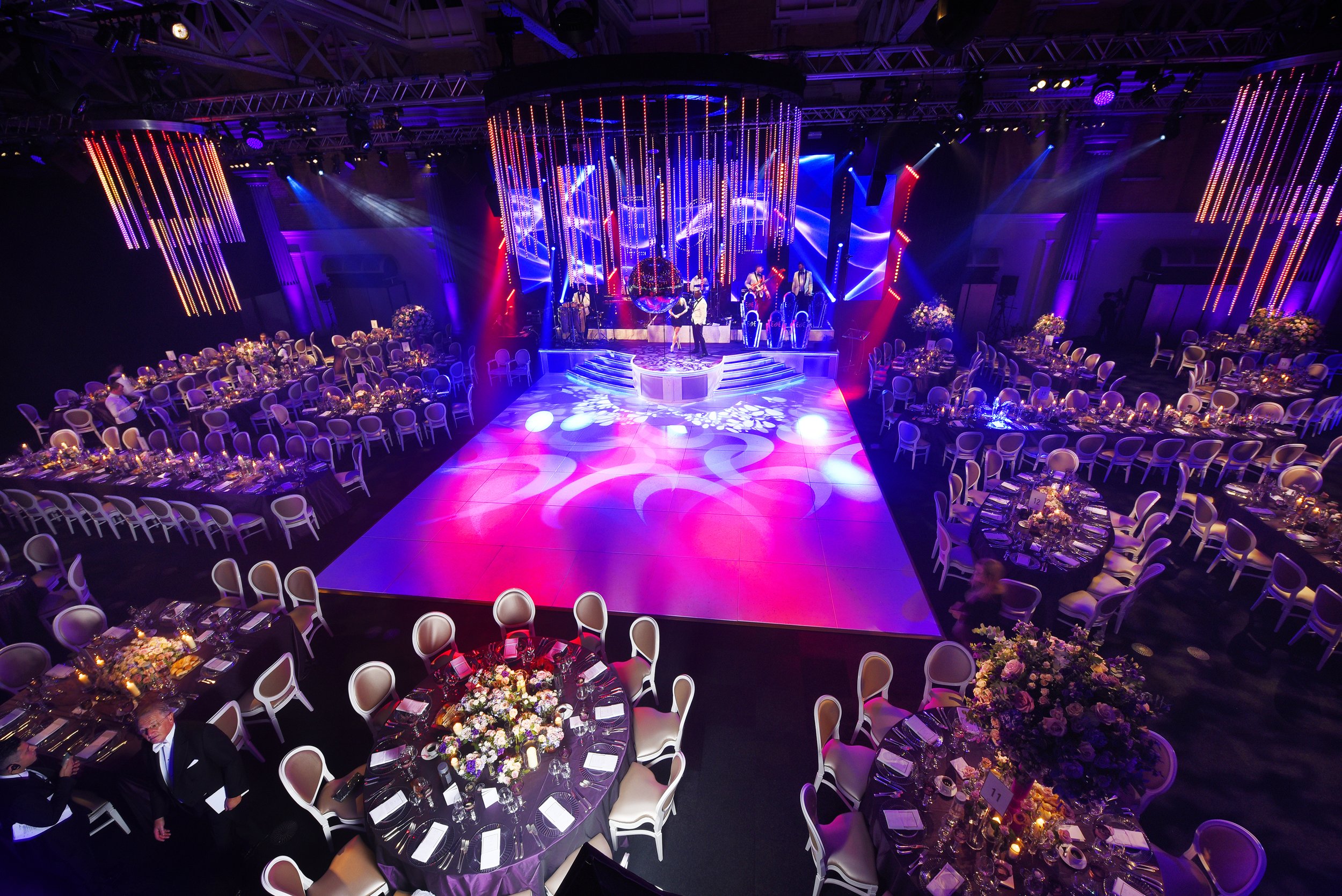 An event hall decorated for a celebration with round tables covered in purple tablecloths, floral centerpieces, and place settings, with a stage set up for live musical performance illuminated with colorful lights and hanging vertical LED strips.