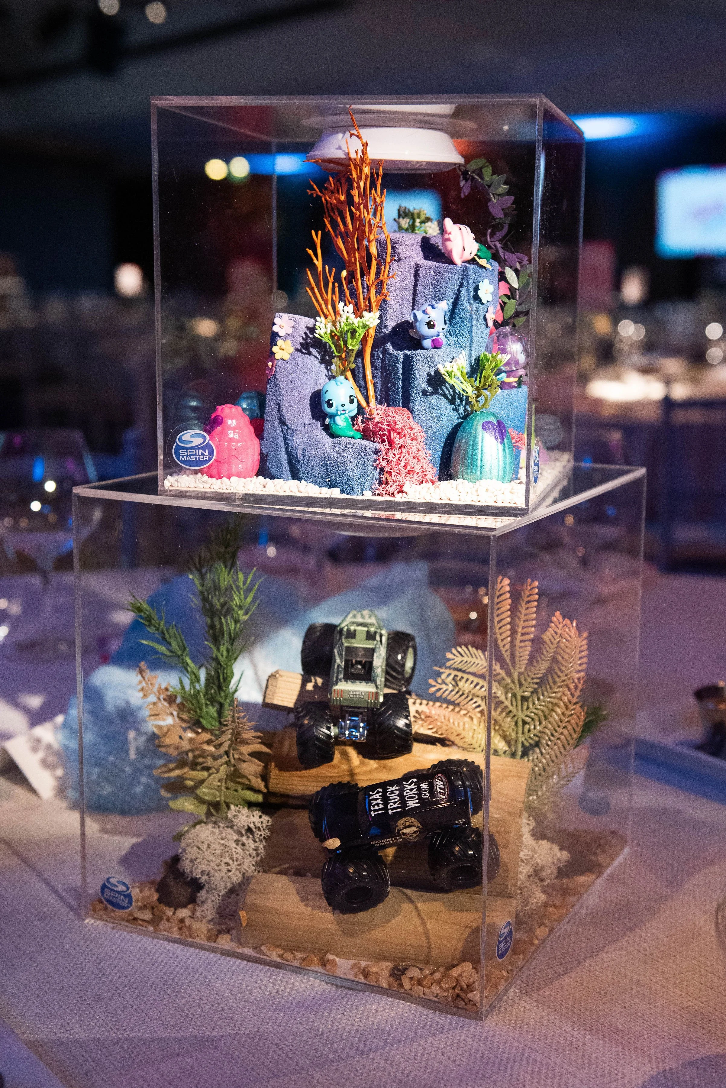 Display of small toys, including a blue animal figure and a pink pig figure, on a tiered platform inside a clear acrylic case with decorative plants and rocks. The background is blurred with some blue and white lights.