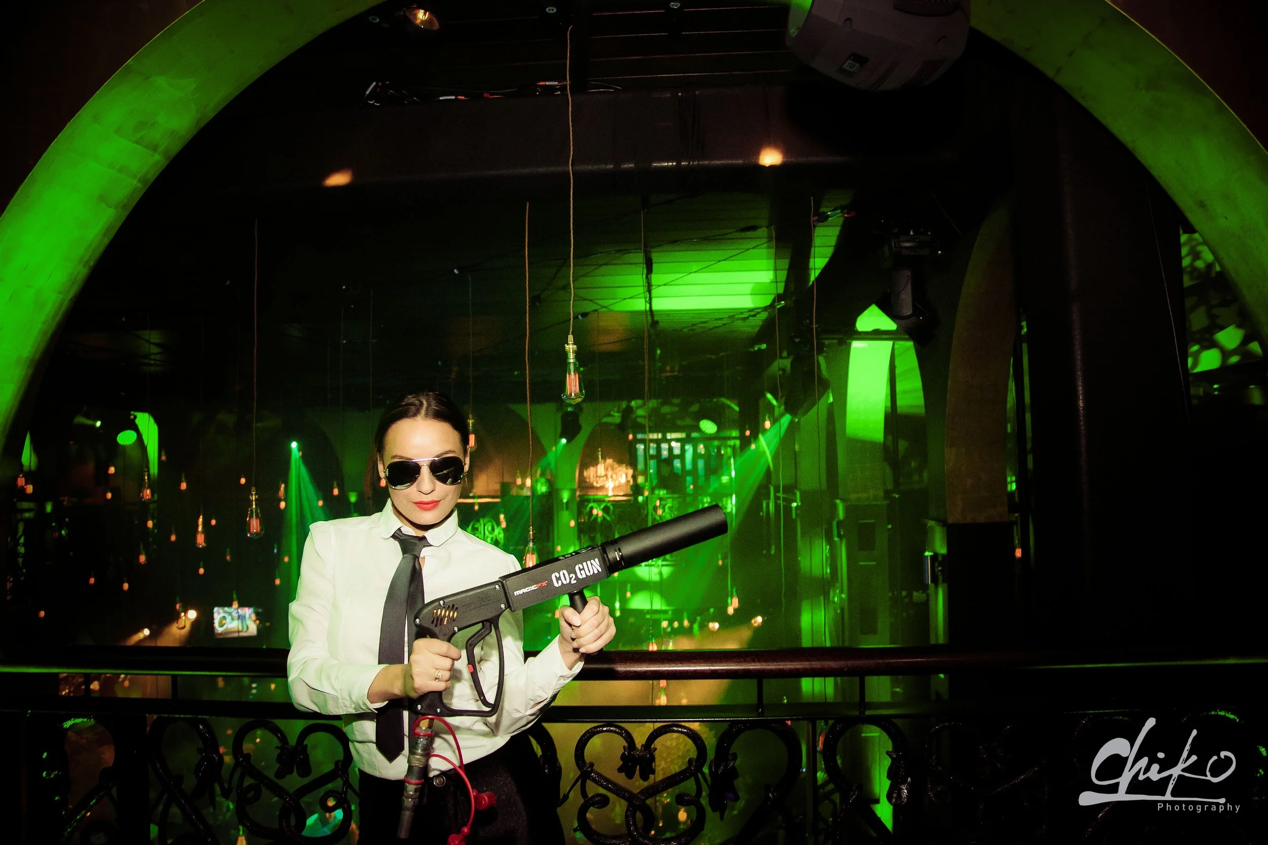 A woman dressed in a white shirt, black tie, and sunglasses holding a CO2 gun in a dimly lit club with green lighting, hanging light bulbs, and a decorative black iron railing.